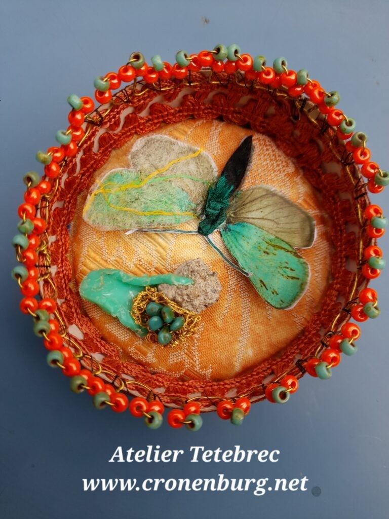 A preciously ornated round cheese box with hand-made perlage and braid in orange and green colour shades. Inside is a green-grey moth, Jordanita subsolana. The wings on one side are hidden under a hand-made gossamer of raw silk, my symbol for vanishing. Once native to Alsace, they are now considered to have disappeared.
A tiny precious element in the same green as the moth is a piece of plastic waste that melted into grey concrete. Glass beads are fixed into a golden crochet "flower".