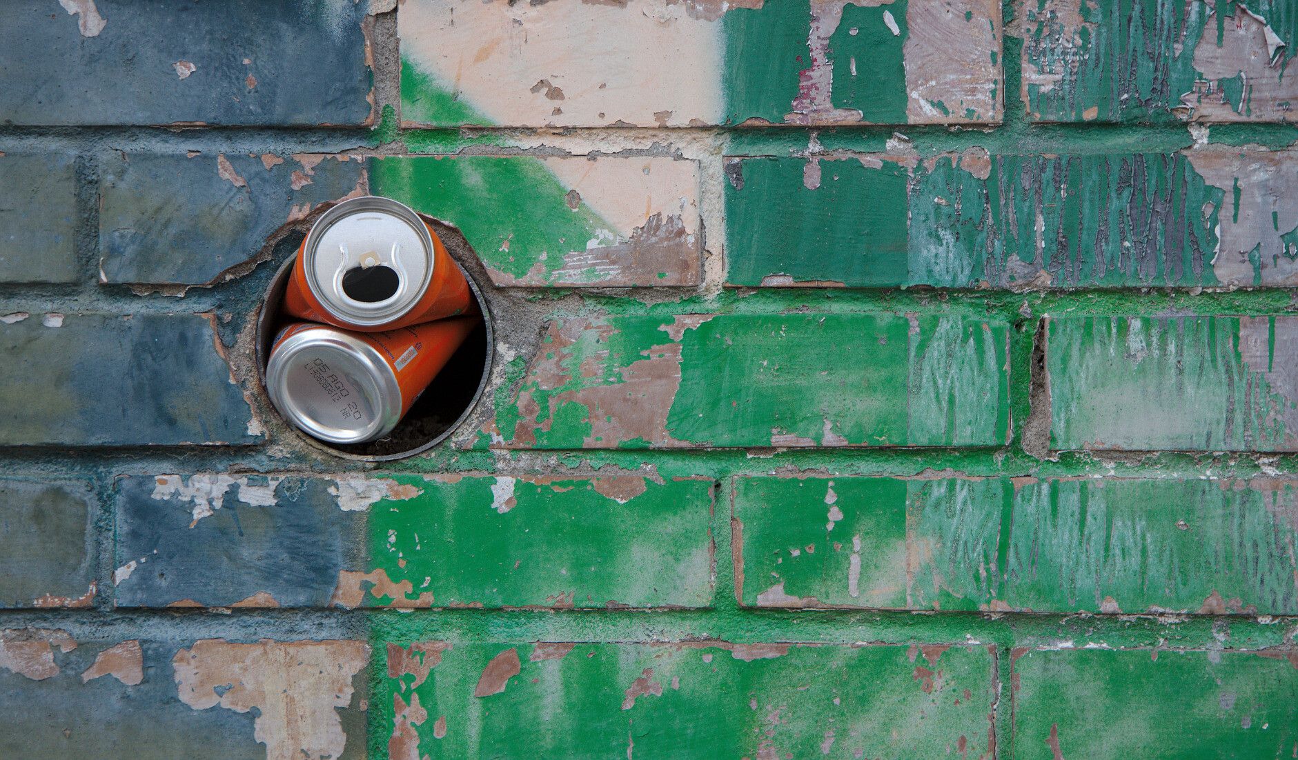 Photo of a wall covered in teal and green graffiti, with a bright orange can of soda stuffed in a hole.
