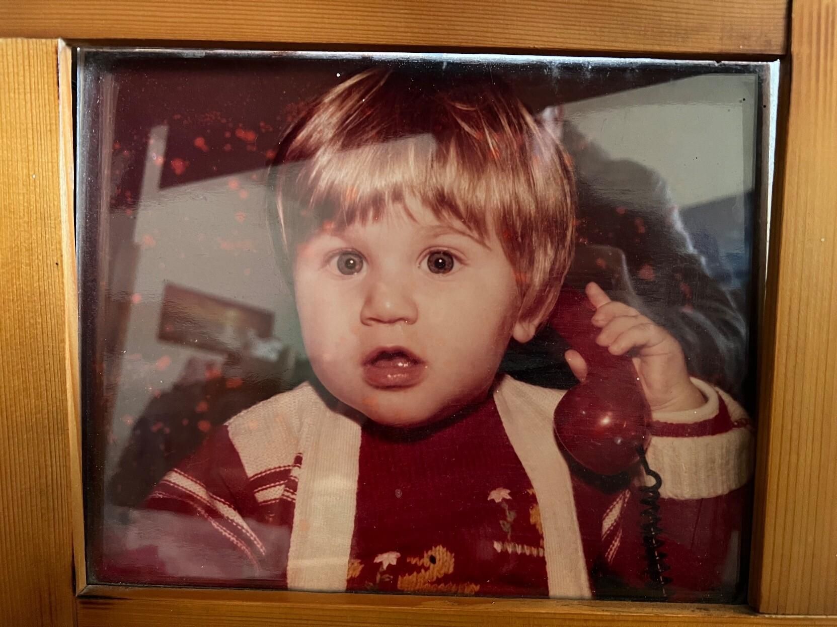 A photo of a photo. I am a young boy with light eyes and blonde hair, wearing a red wool sweater and holding a red toy phone in my hand. This photo will always remind me of my grandparents' living room and their house.