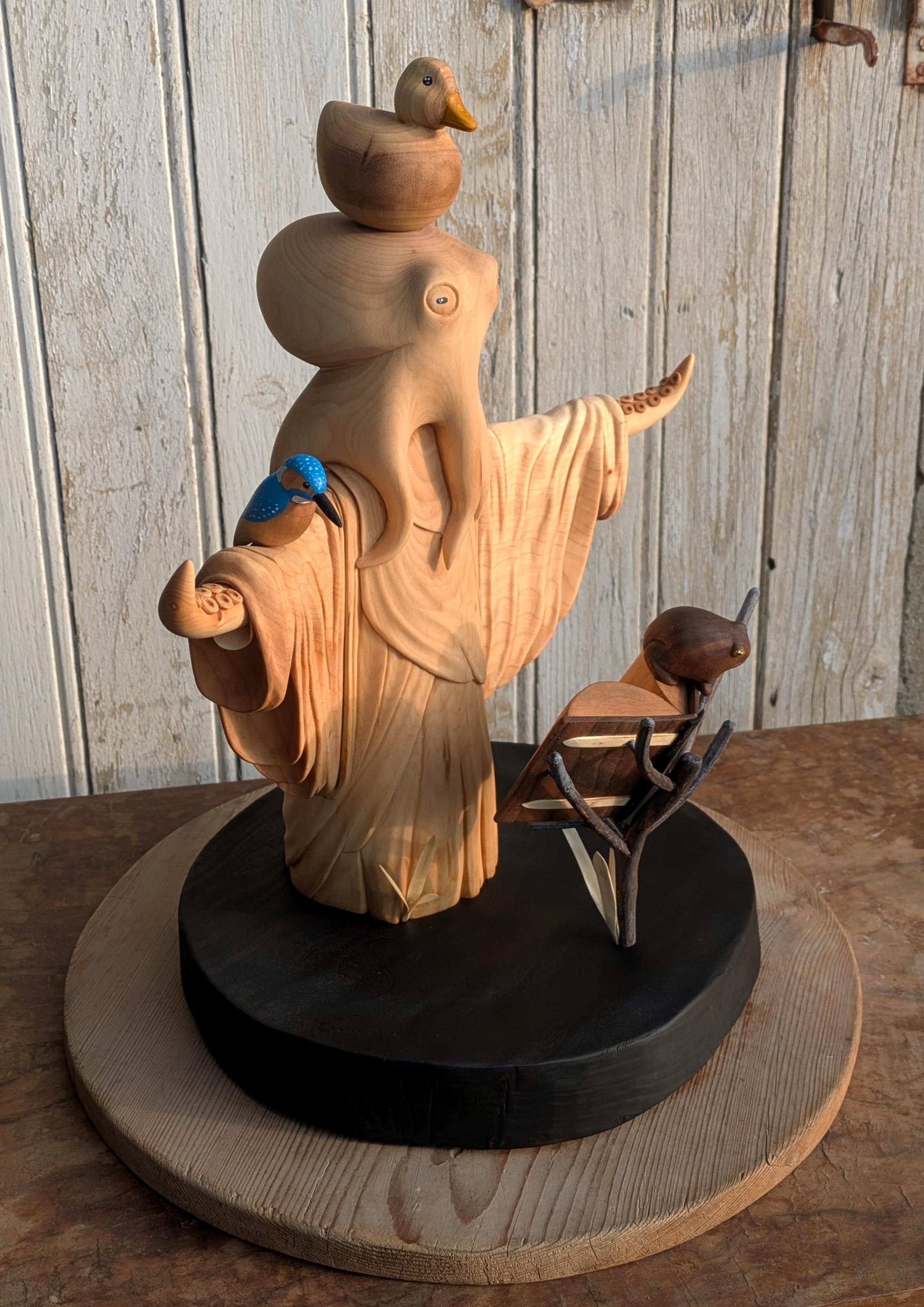 Woodcarved humanoid octopus wearing a robe, arms wide open, in front of a book standing on branches. On the book, a frog. On the right arm, a kinfisher, and on the top of the head of the octopus, a round duck. The base is burn, on that angle it is not really obvious but is water with a slight wave form arround the octopus. On the background, an old door with old white paint.
