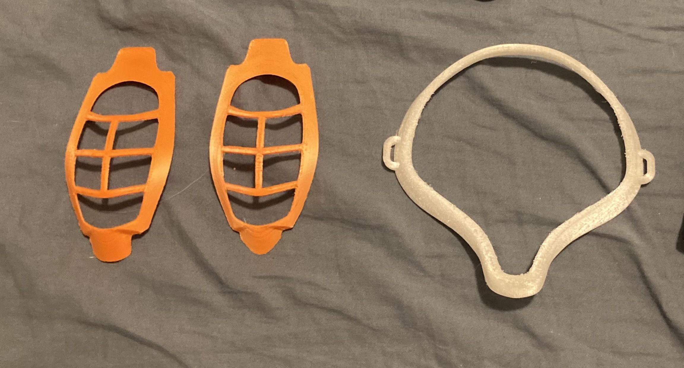 3d printed accessories for cloth breathing masks. Two orange mask inserts on the left, which are for preventing the mask from collapsing when breathing in. A white mask frame on the right, which goes around the outside of the mask for improving the air seal between mask and skin.