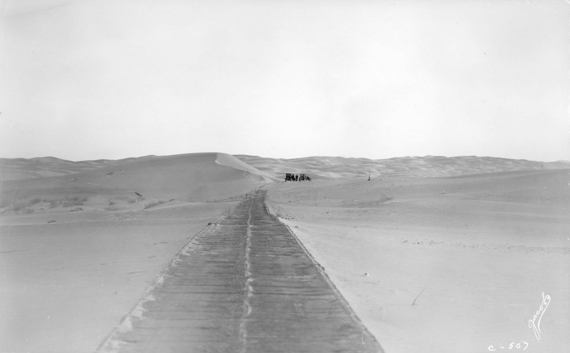 From source:
Summary
View of plank road in sand dunes near Yuma (Yuma County), Arizona. Cars are in the distance.
Notes
Photographer's byline and "C-507" reproduced in photoprint. Title penciled on verso with "Presented to Jack McKincrurie (?) by John Rogers at Yuma Arizona Aug. 21st 1926 - thermometer 106.  We crossed planked road Aug 19. This road discontinued and cement highway opened Aug 20th 1924. This range is 6 miles across and several miles long and shift first into one position and then another like the snow in our eastern blizzards. The road at times has disappeared under 20 or 30 ft of sand for a mile or more. Then a new section of road is built to fill in the missing link and its grade maintained among the sand dunes," and "Jack McKincrurie (?), Jan 20, 1985, Glenn & Hazel Shomaker gave me this picture, George A. McClellan."
