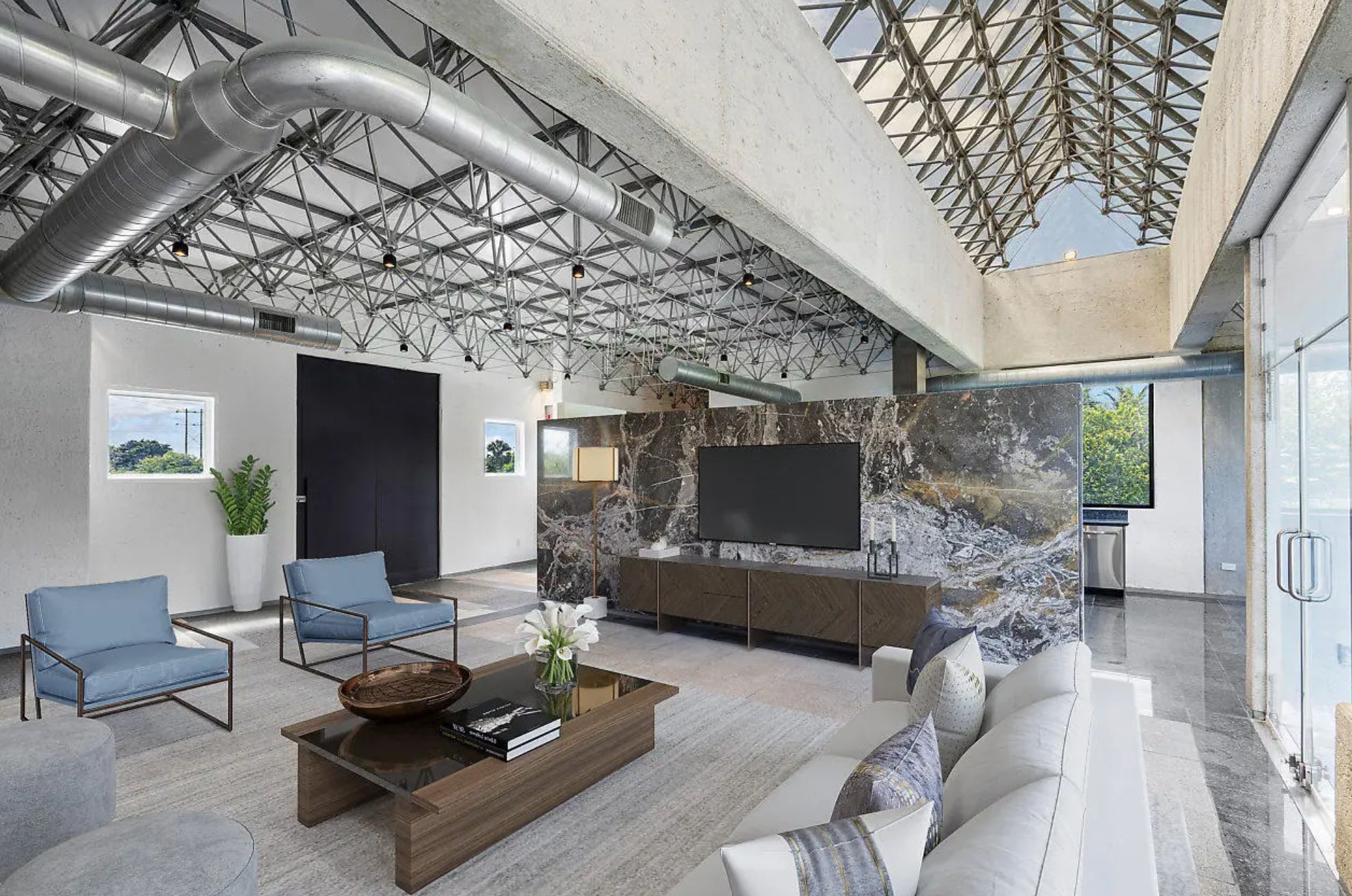 An open plan living area with kitchen visible a bit behind a marble wall. The ceiling is unfinished and shows a latticework of metal beams and exposed HVAC system. The furniture looks AI generated. 