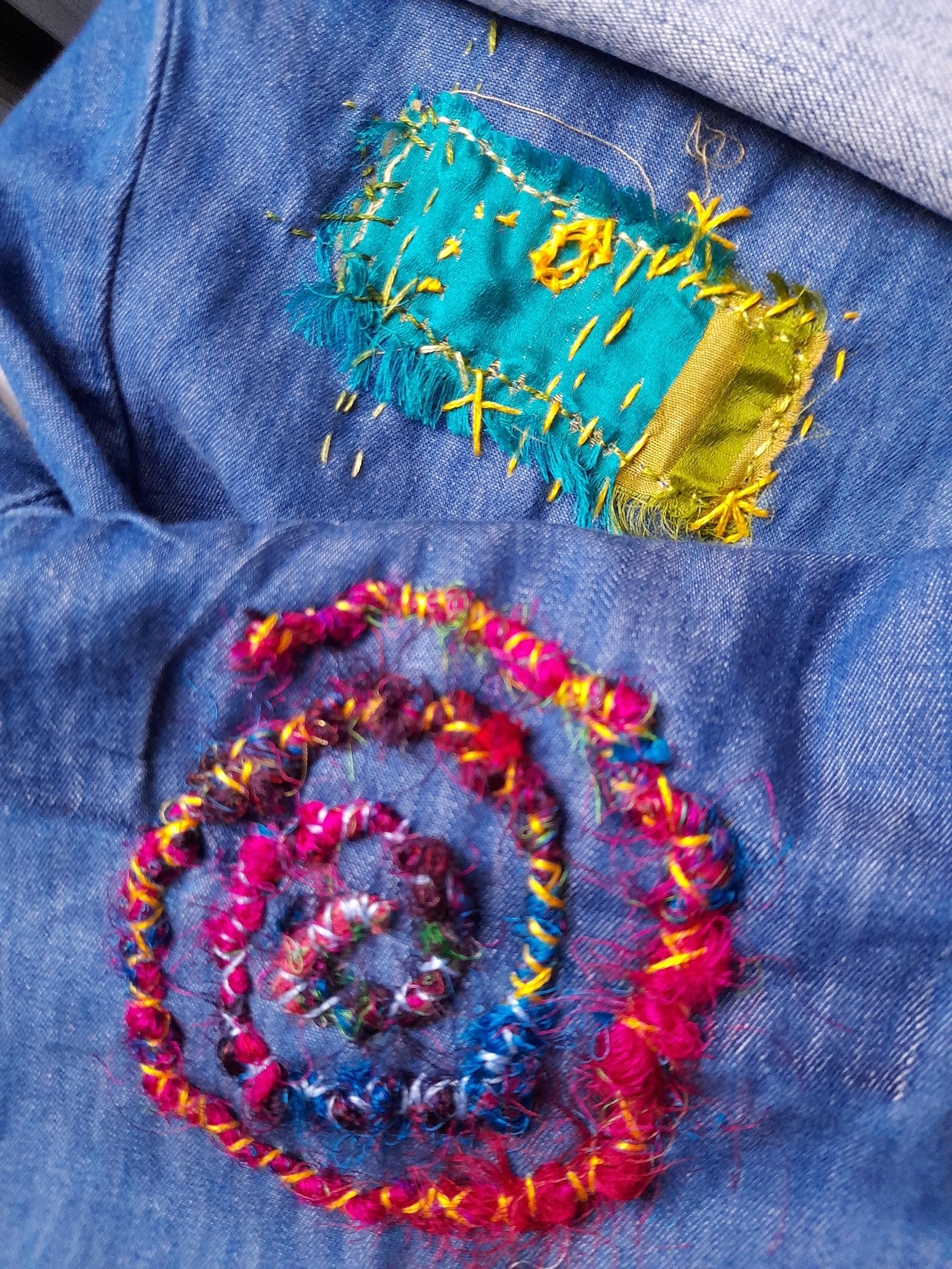 Visible mending on jeans fabric: a pink-purple-blue spiral of silk threads fixed with yellow stitching. And a rectangular silk patch with turquoise and golden fabric, fixed with intuitive embroidery in the same yellow.