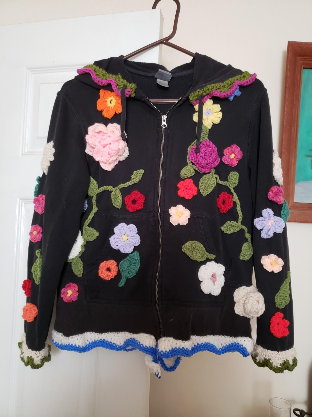 The front of a black hoodie that has multicolor crocheted flowers, leaves, and vines on it as well as crocheted trim on the hood, sleeves, and bottom.