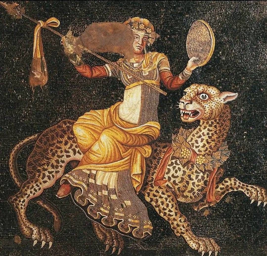 Image description: Dionysus rides side-saddle on the back of a panther with a ribboned thyrsus (a pine-cone tipped staff) in one hand and a tambourine in the other. Dionysus wears flowing robes and a crown of foliage and flowers.