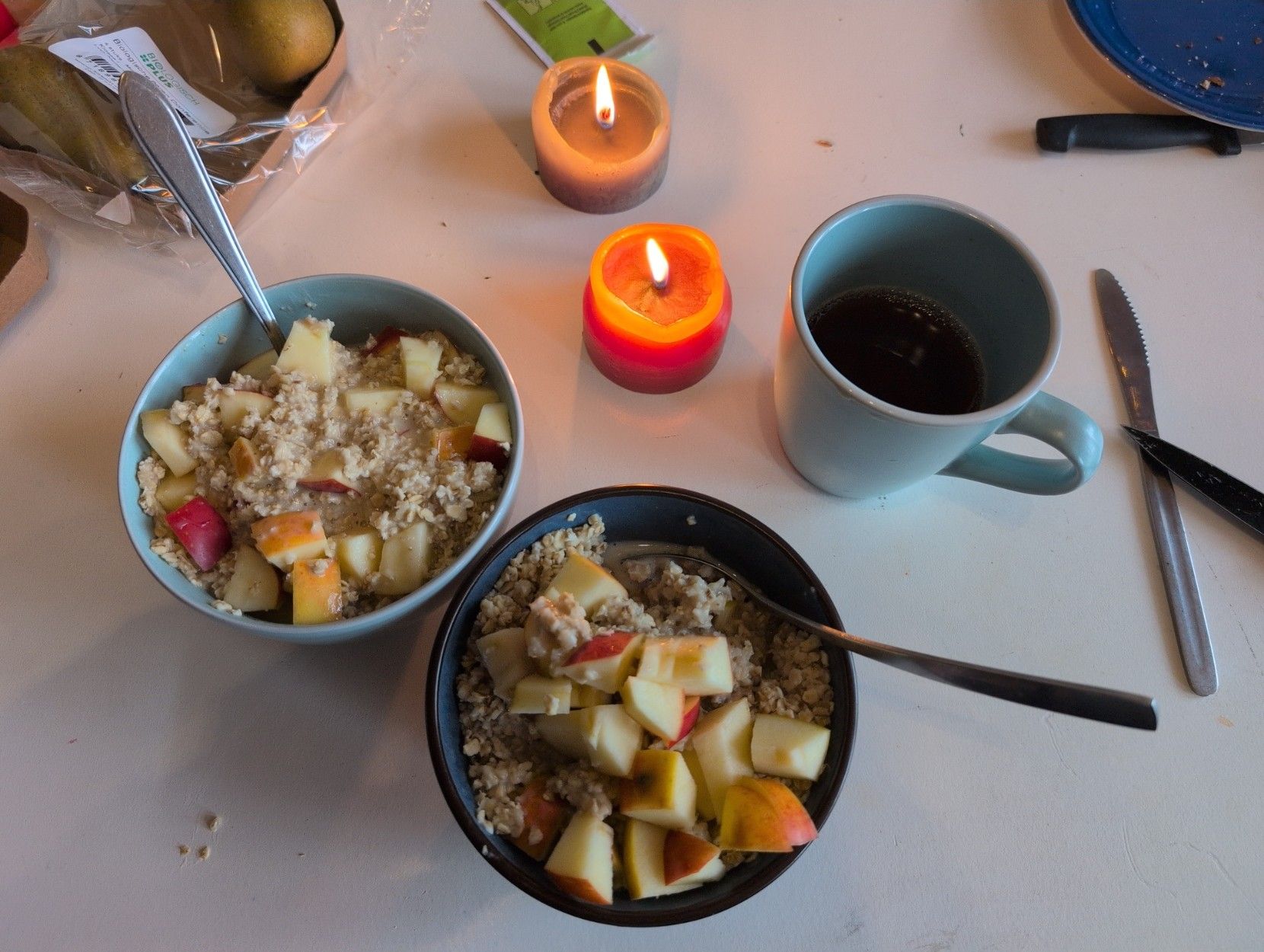 Photo of two bowls of porridge with pieces of apple, a cup of tea, two candles.