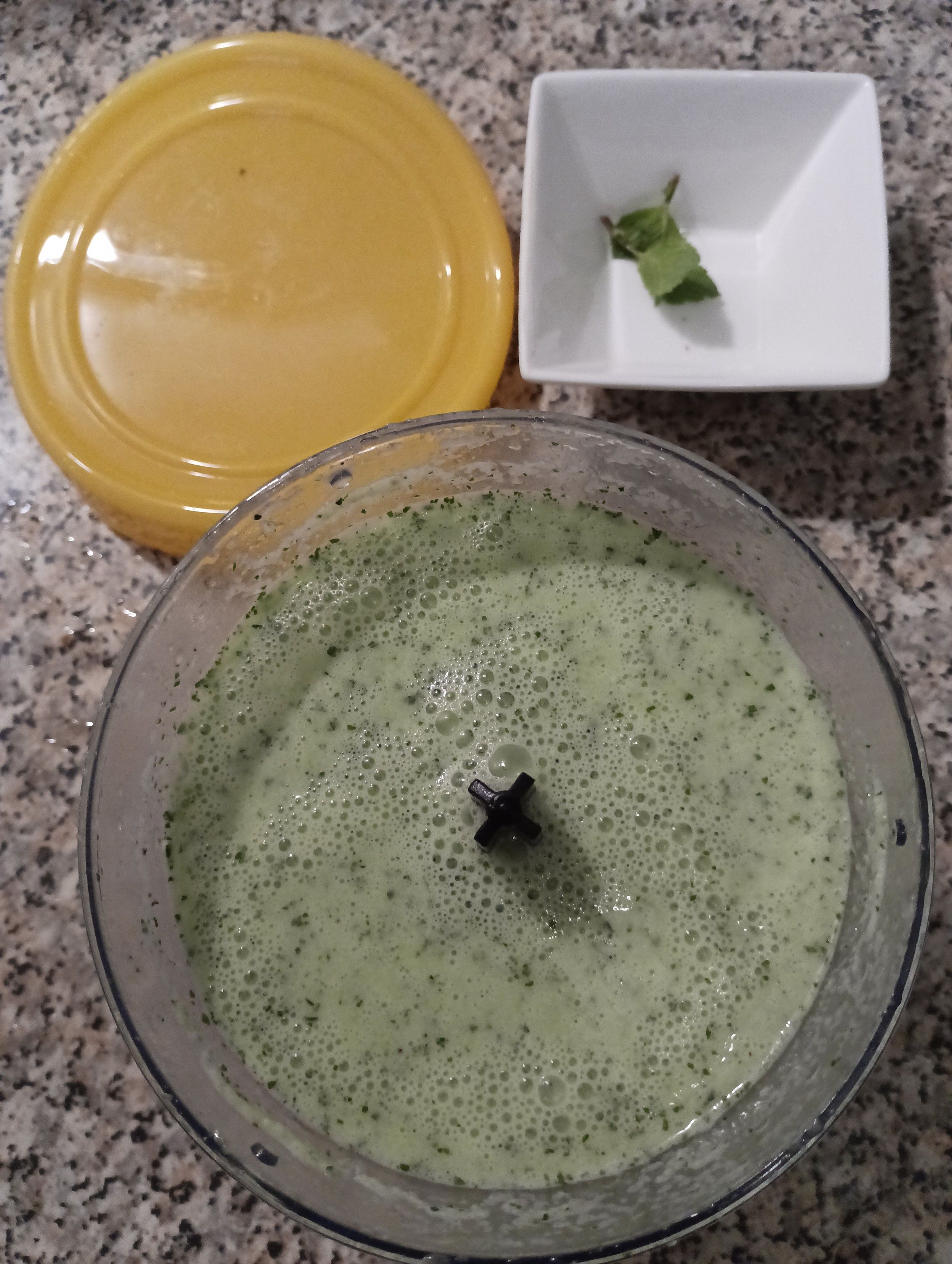 Foto do mesmo vaso de triturador con todos os ingredientes xa triturados, xusto antes de poñelo a arrefriar no frigorífico. Á beira a tapa de silicona para o vaso e mais unha cunquiña con puntas de hortelá coas que decorar após as cuncas de servizo.