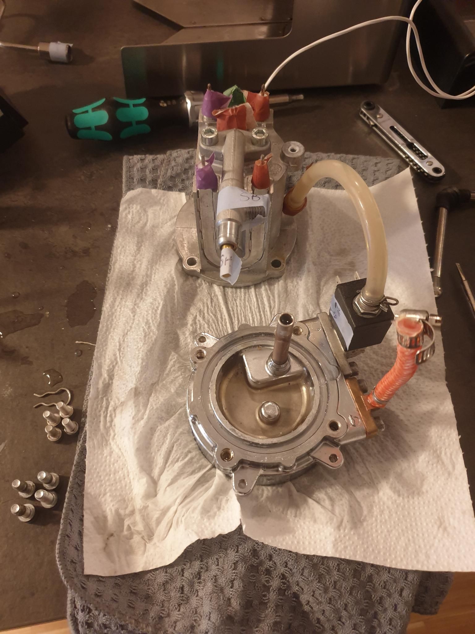 The disassembled boiler rests on top of a soggy paper towel and kitchen towel. Some of the tubes and most of the wires are removed from it, minus one wire that looked like some special connection I don't know how to correctly remove or reattach. Nine screws are lined up to the side.