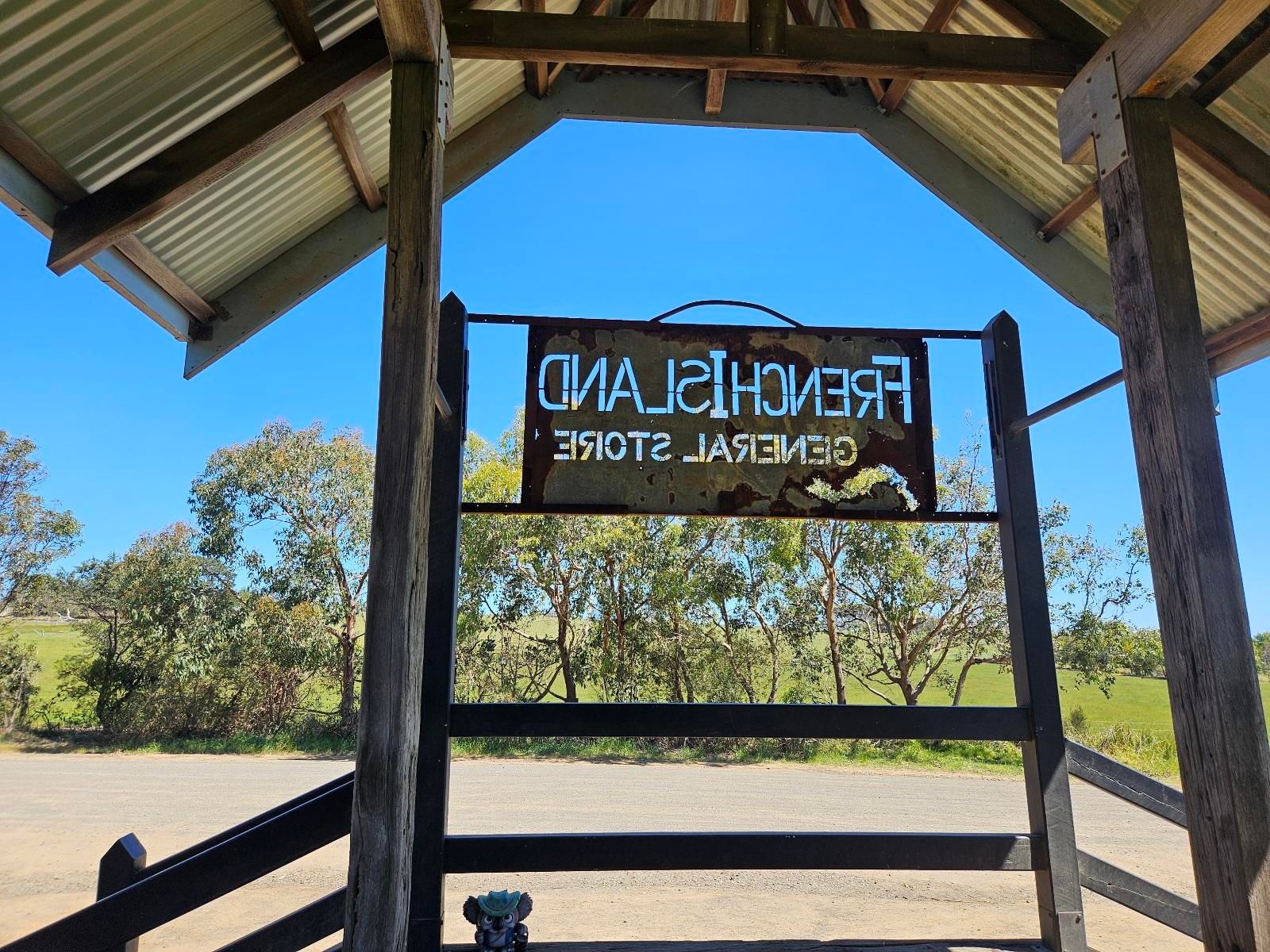 The partly rusted out sign at the French Island General Store, viewed from behind so that it's backwards. You can see a dusty track in front, and a eucalypt-bordered field beyond that. Above, clear blue skies and the merciless sun.