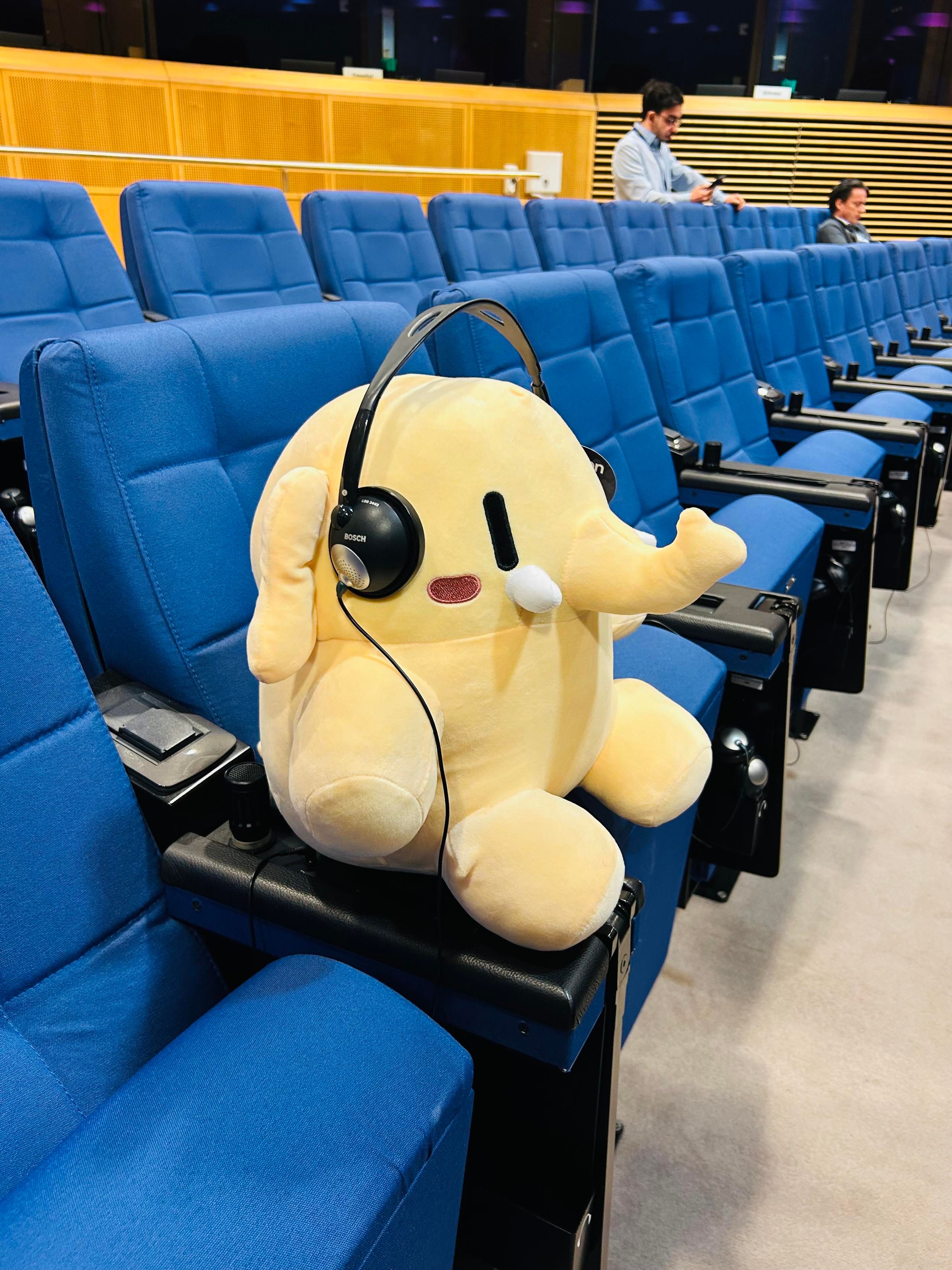 The mastodon plushie (a little elephant) sits on a blue chair in a row of blue chairs, wearing a translation headset 