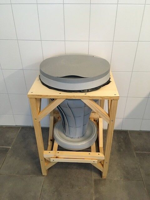 A funnel-shaped plastic bin, with the narrow end pointing down, suspended in a wooden frame. The funnel consists of flaps that have been bent and woven together, which makes it look somewhat like a tornado. There is a lid on top. A plastic dish is pressed against the underside of the funnel.