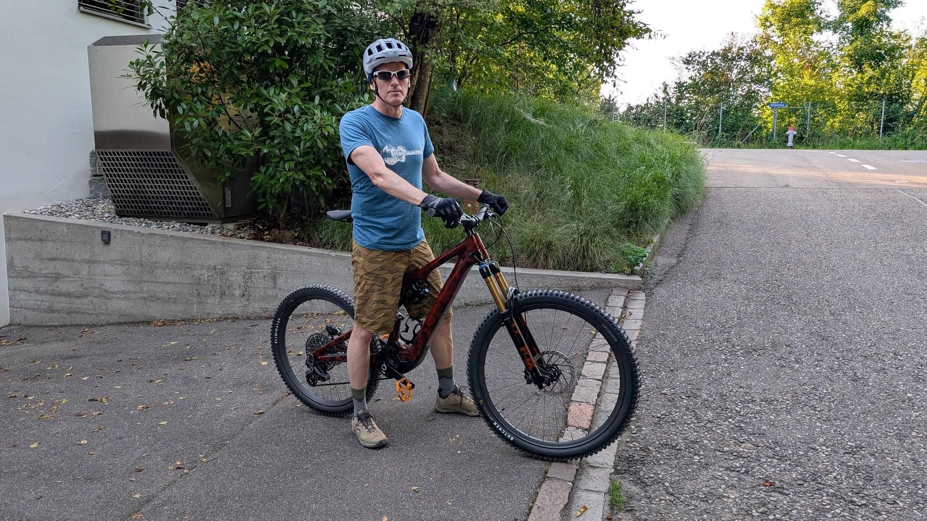 Photo of a man with his brand-new eBike. He's wearing his MTB outfit, helmet, and tinted glasses. Behind him, greenery and the wall of a house.