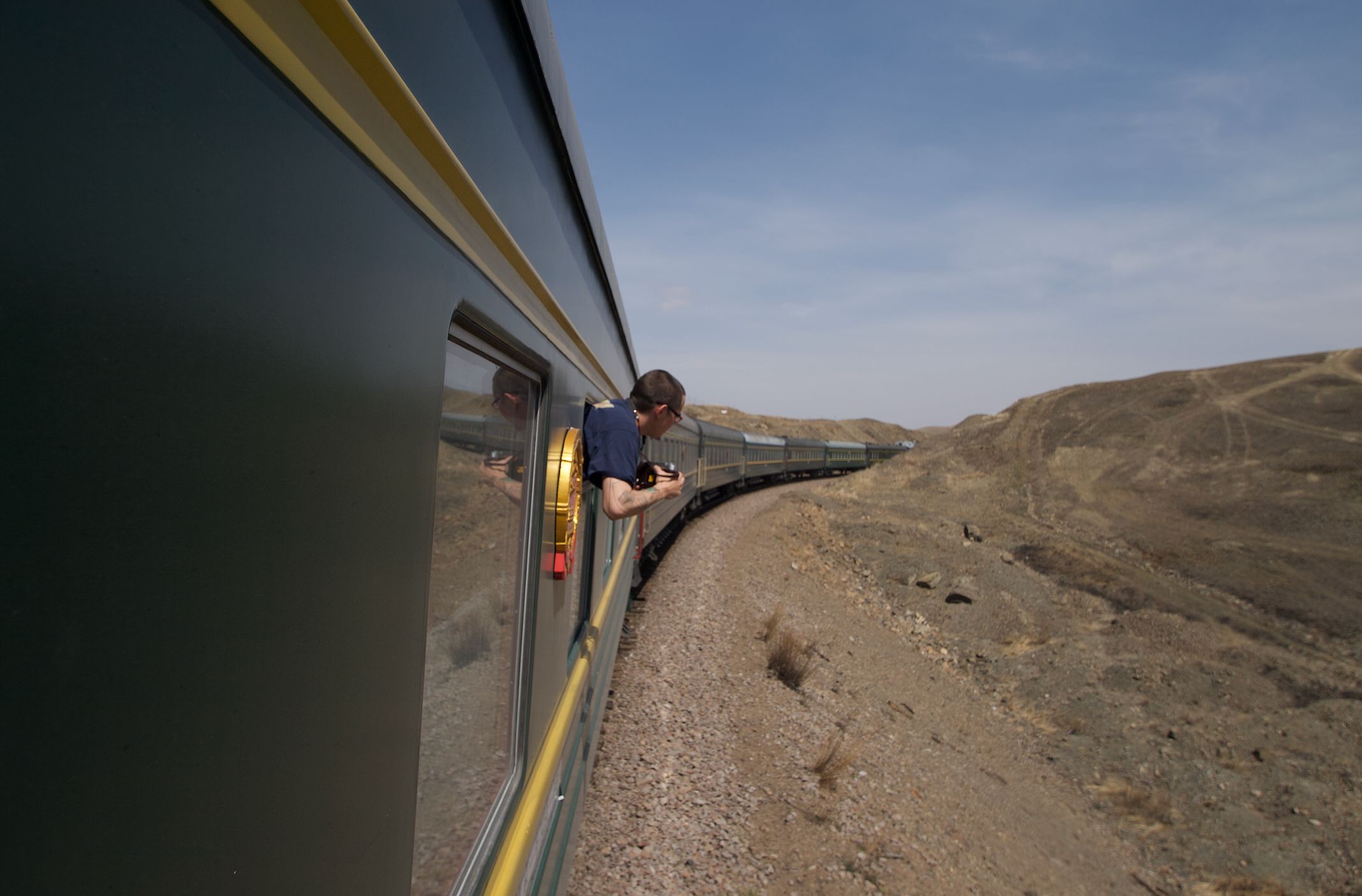 on the left, the side of a green train with yellow trim can be seen. a man leans out of it, with his camera from a window ahead. in the distance and on the right, a desert landscape is seen, with a scrubby brown-green hill on the far right., it all sits under a hazy white blue sky