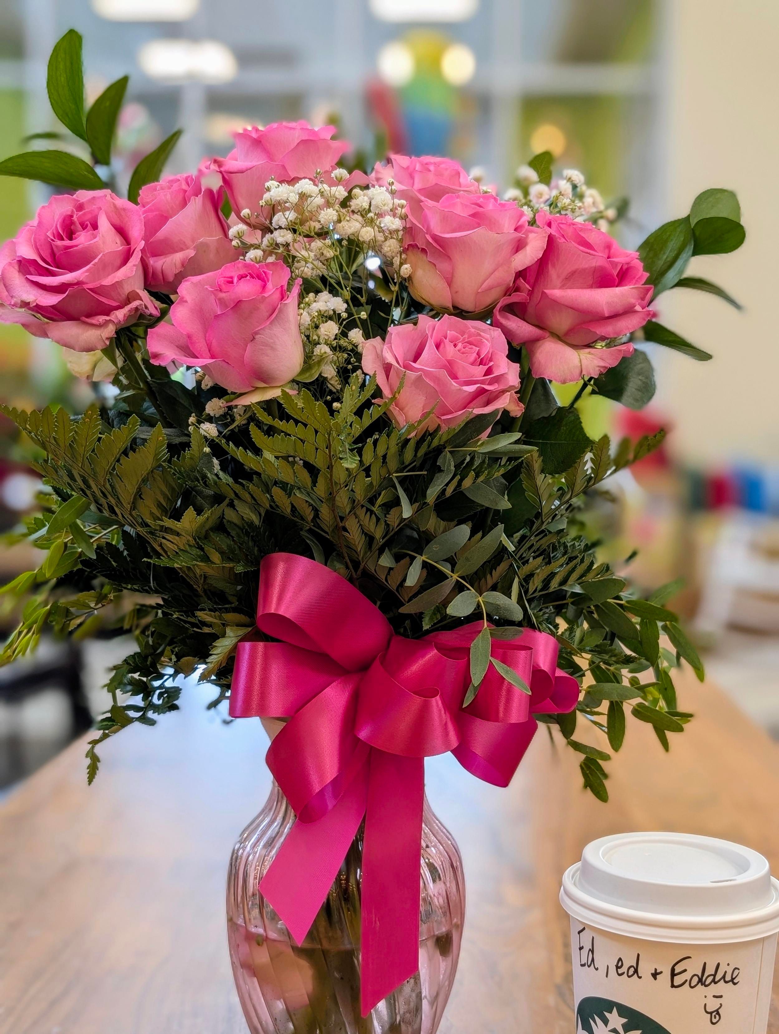 vase of pink roses sits next to a carryout coffee cup on a long wooden table. tall, out of focus windows to the outside in the background. 