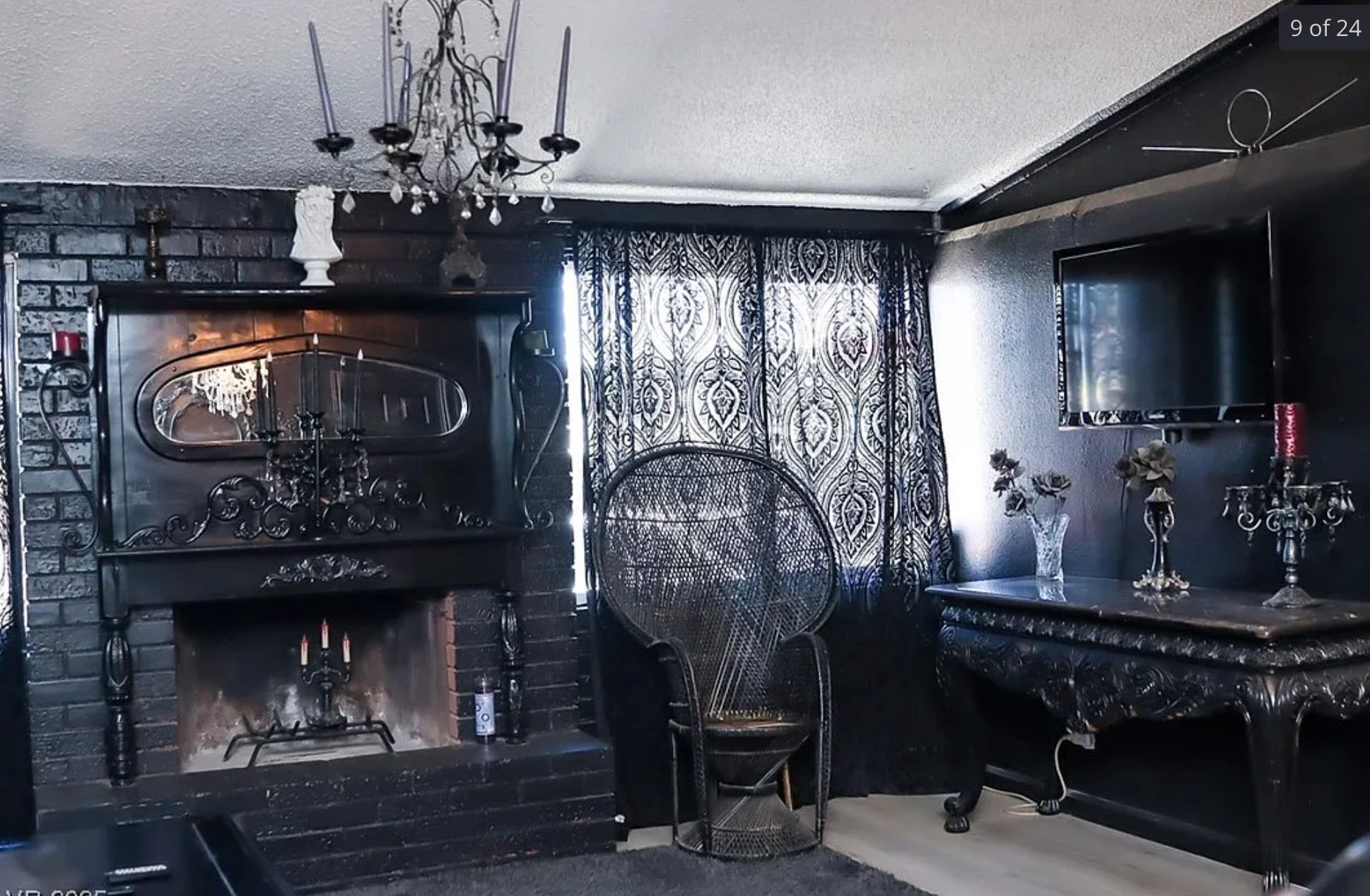 A living room that is entirely black except for the ceiling. The fireplace is black, the chairs and curtains are black, the chandelier is black, and the side table is black. 