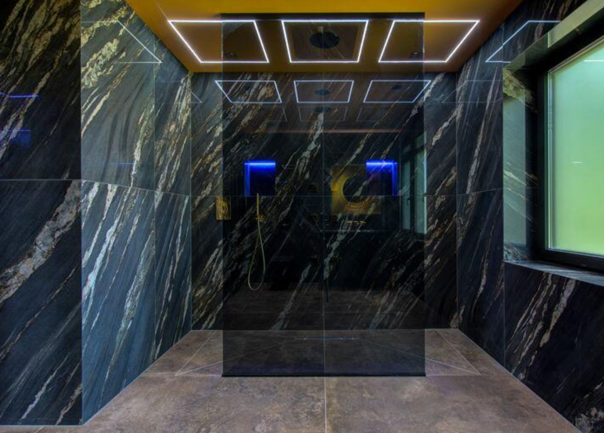 A bathroom entirely clad in dark marble and what appears to be a shower in the middle. The glass is reflective and the ceiling has a series of squares that are lit around the edges. It is very hard to explain and is very confusing. 