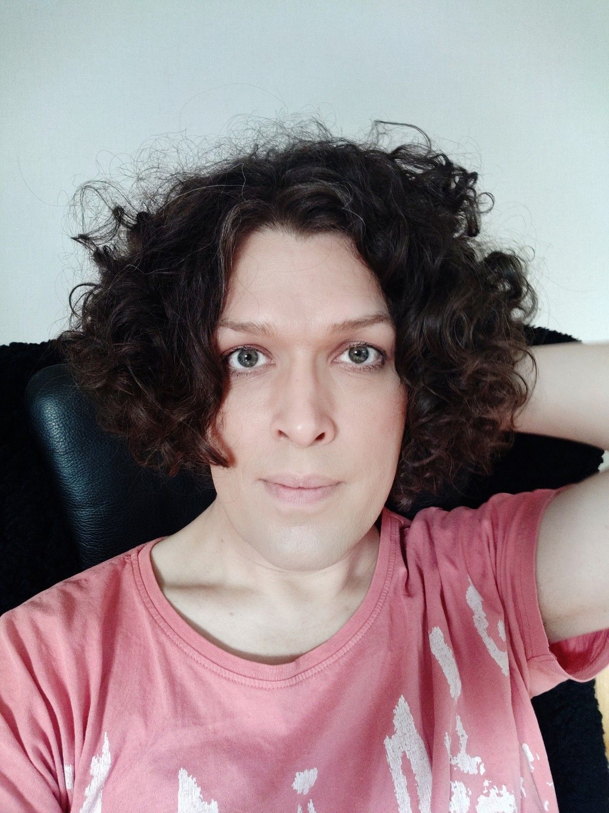 Me with my left hand behind my neck, sitting in a black armchair in a pink and white t-shirt. I'm smiling a little and looking pretty okay.