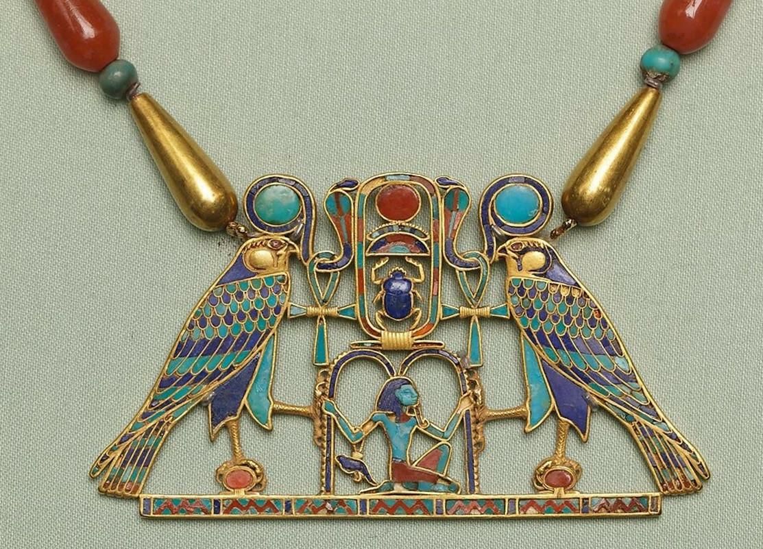 An incredibly intricate necklace in red, blue, purple an gold of two birds facing each other, a man kneeling on the ground beneath them.