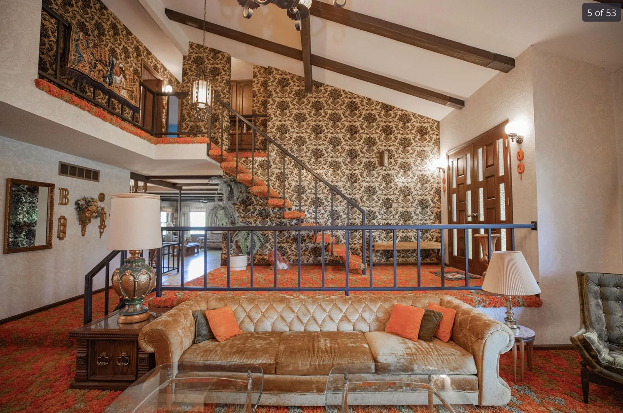 A view of an open plan living area and stairs going up to the 2nd floor in the background. The style is very 1970s with orange carpeting and brown patterned wallpaper and a gold velour couch. 