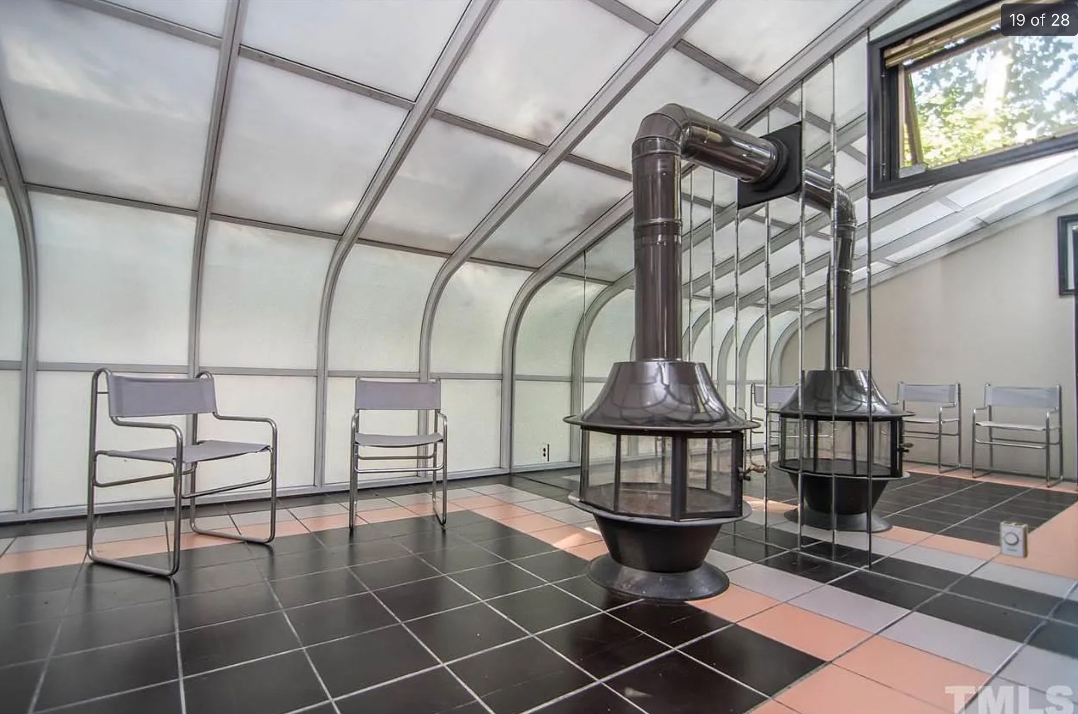 A room with 2 chairs that look like they belong in an office, a tiled floor, and what looks like a round stove heater. The room is otherwise empty and the wall is mirrored. There is frosted glass along one curved wall. 
