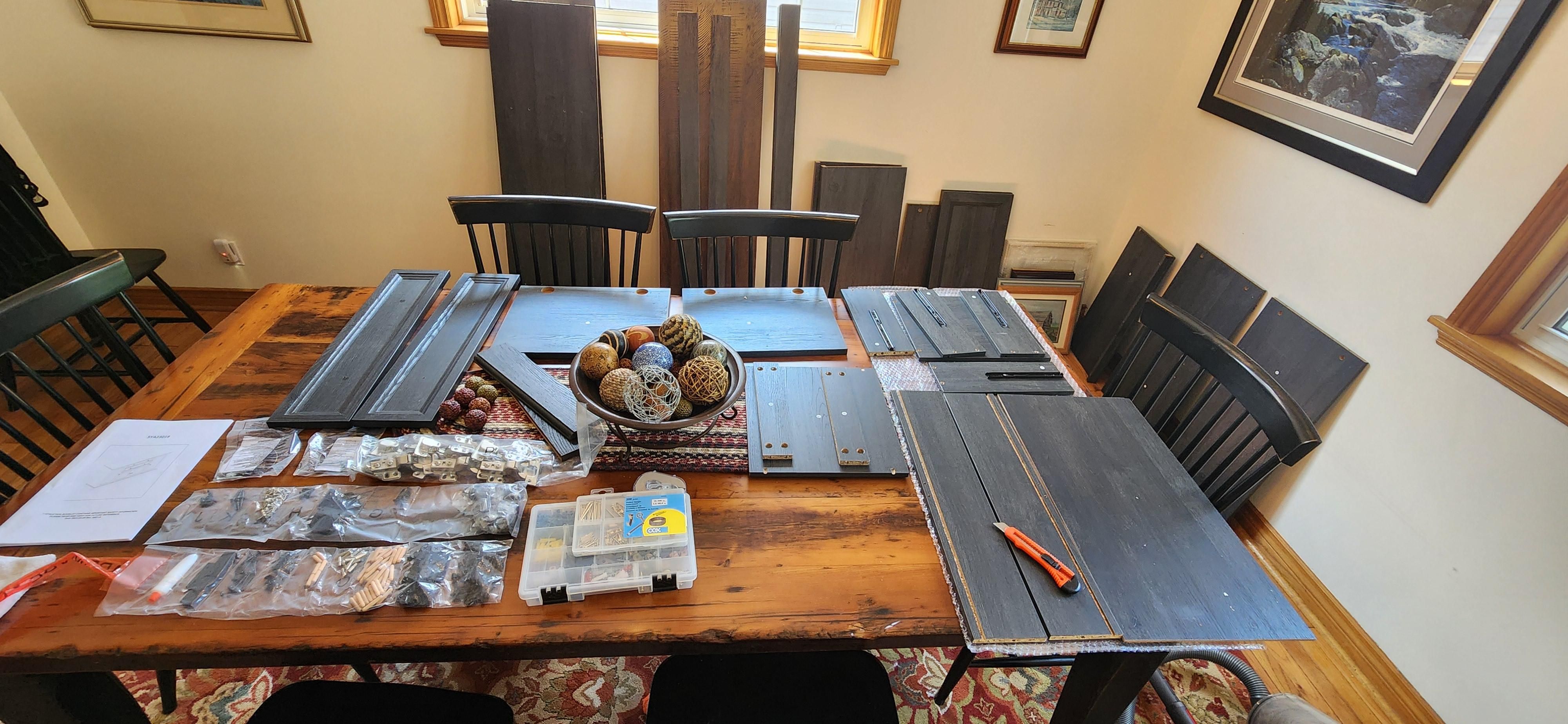 A picture of a dining room with 40+ pieces of a disassembled sideboard spread around the room. Some are laid across our table and larger pieces are leaning against the wall. In the bottom left corner, there are 5 plastic bags full of different types of hardware needed to assemble the furniture.