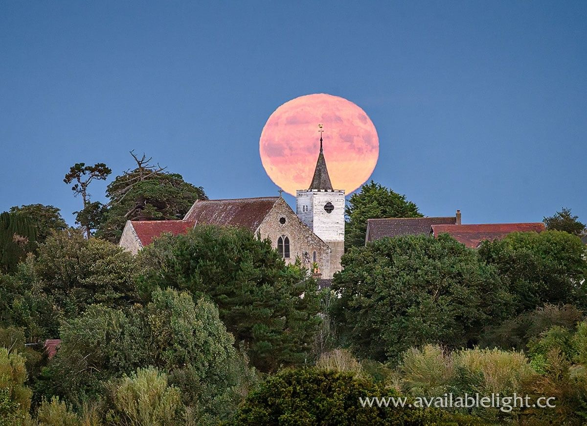 A slightly tatty looking church spire with a huge moon behind it. It's dusk and the sky is a clear deep blue, the moon very bright. The church is surrounded with trees.