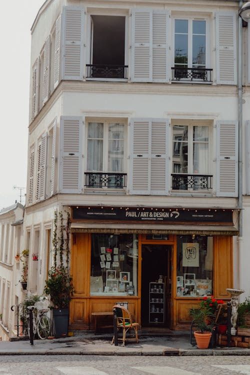 A white three-floor building on the corner of a cobbled street, with white wooden shuttered windows that have short black iron railings in front of them. The front of the lower floor that opens onto the street is wood-faced, with some chairs and potted lants outside, and a bike leaning against the corner. 
