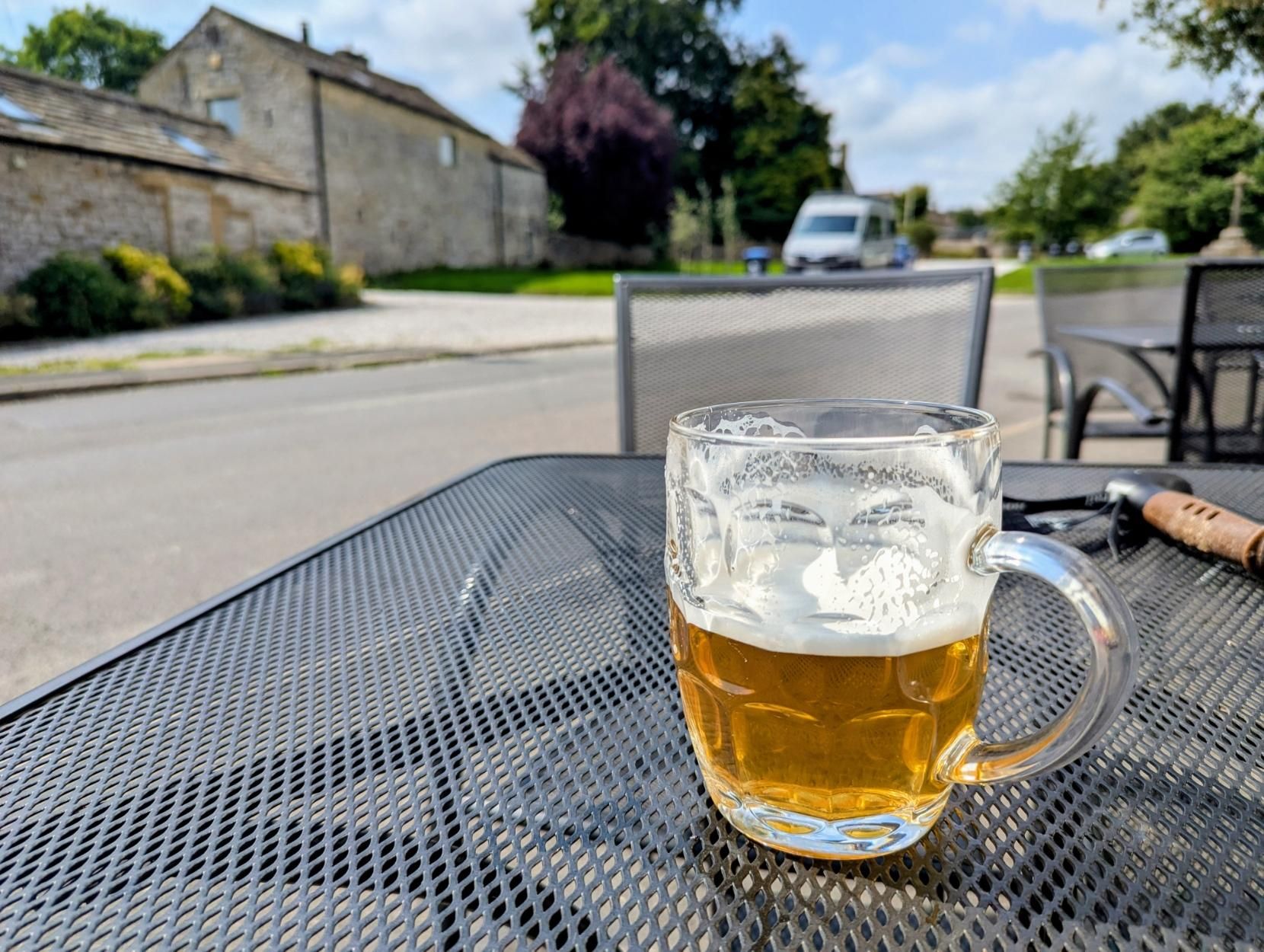 A half full pint dimple glass on an outside table. A road and old stone buildings are visible behind.