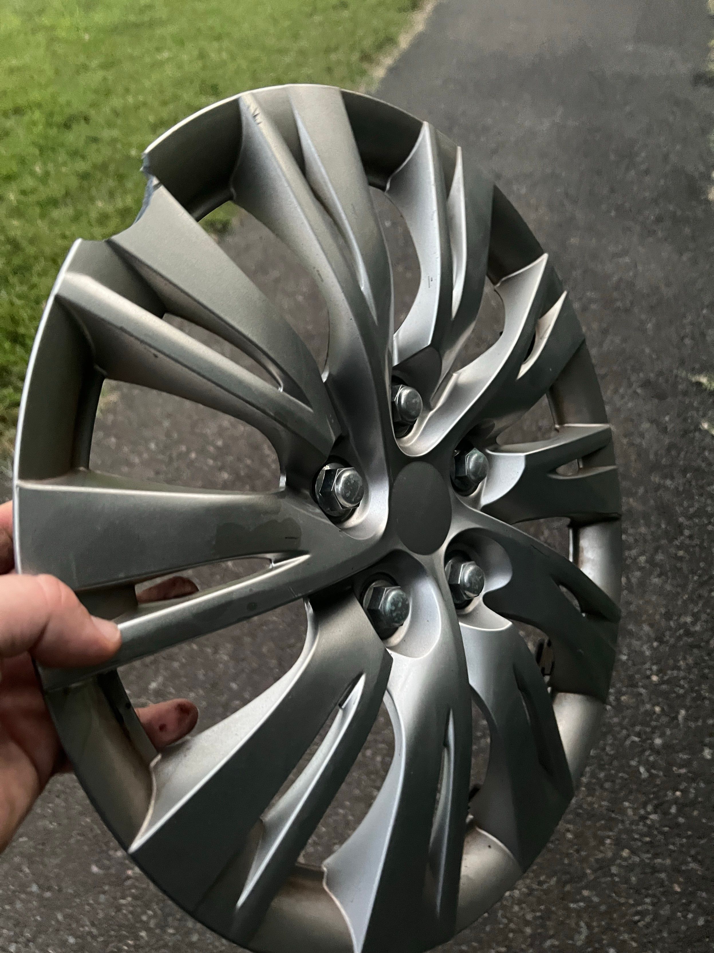 Photo of a hand holding a silver wheel cover. It looks like there are 5 lugnuts. But there aren’t. It’s a solid piece of plastic. 