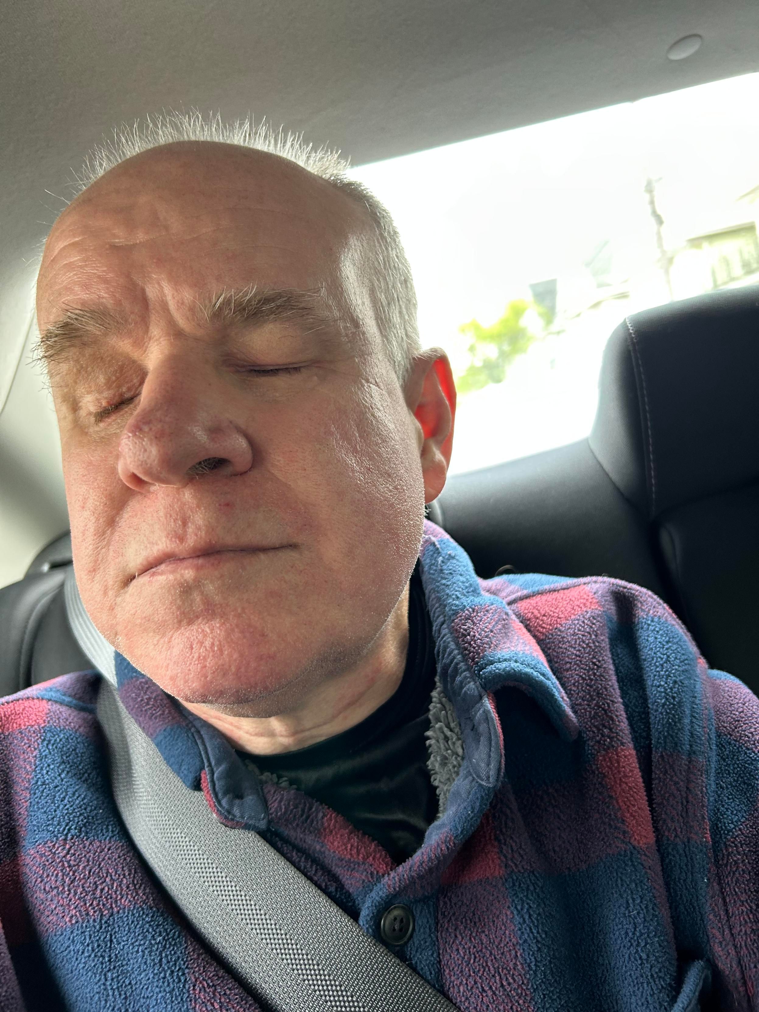 Be My AI: A middle-aged man with short, light-colored hair is sitting in the backseat of a car. He is wearing a blue and red plaid fleece jacket over a black shirt. His eyes are closed, and he appears to be resting or asleep. The seatbelt is fastened across his chest. The background shows a bright window with some greenery and a hint of buildings outside.