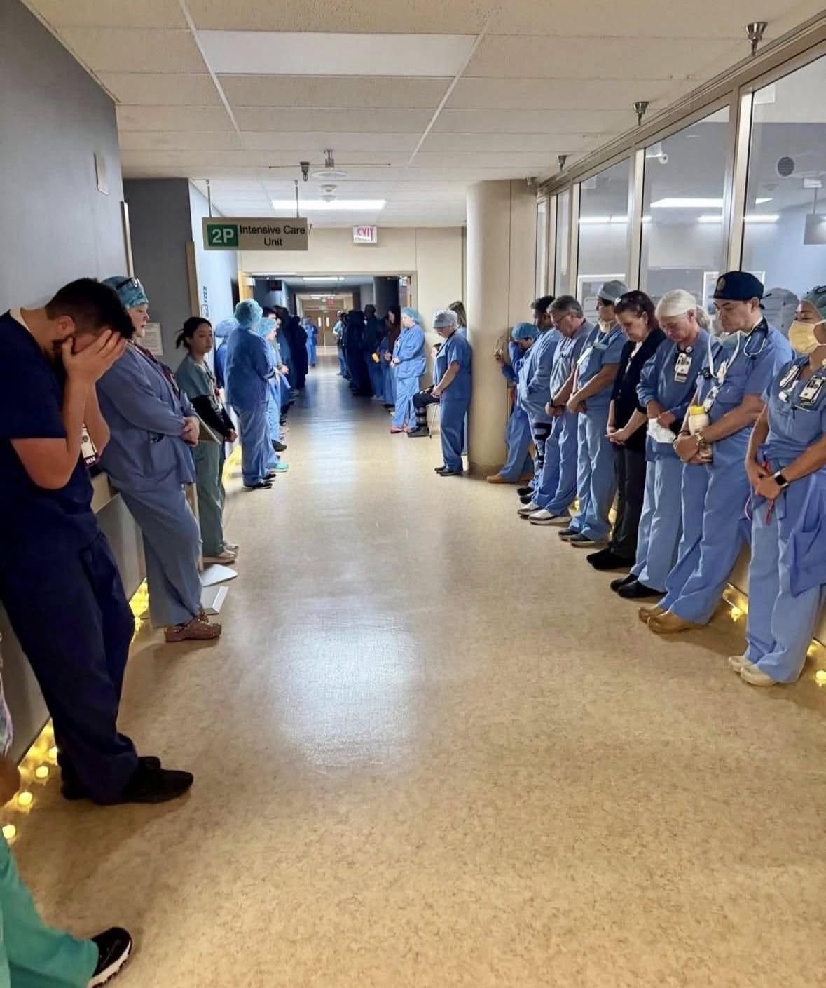 A hospital hallway with a couple dozen healthcare workers lined up along the walls with heads bowed. Some appear to be crying, one has his hands over his face.