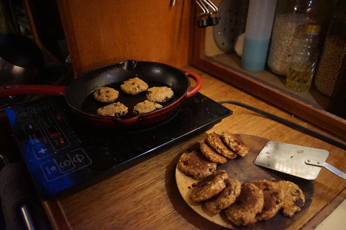 5 cookies cooking in a red cast iron pan over an induction plate. Besides the induction plate is a plate full of cooked cookies, and a spatula.