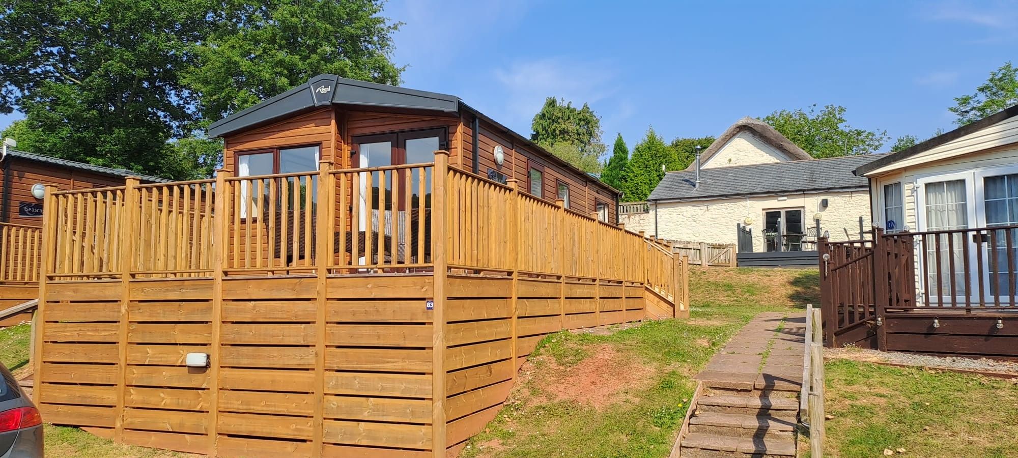 Ein Stativ Caravan der aufgestellt ist. Er steht an einem Hang und das Foto ist schräg von unten aufgenommen. Zu sehen ist eine lange Seite und eine kurze Seite. Der Caravan ist rundum von einem Holzzaun und einer Terrasse aus Holz eingefasst. Er sieht aus wie ein kleines richtiges Häuschen.