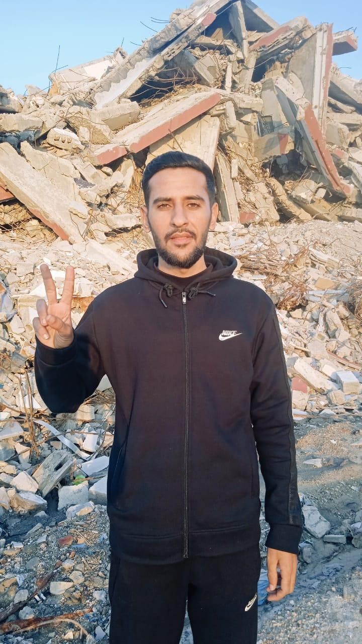 Ahmad devant des décombres en veste noire et faisant le signe v de la main droite