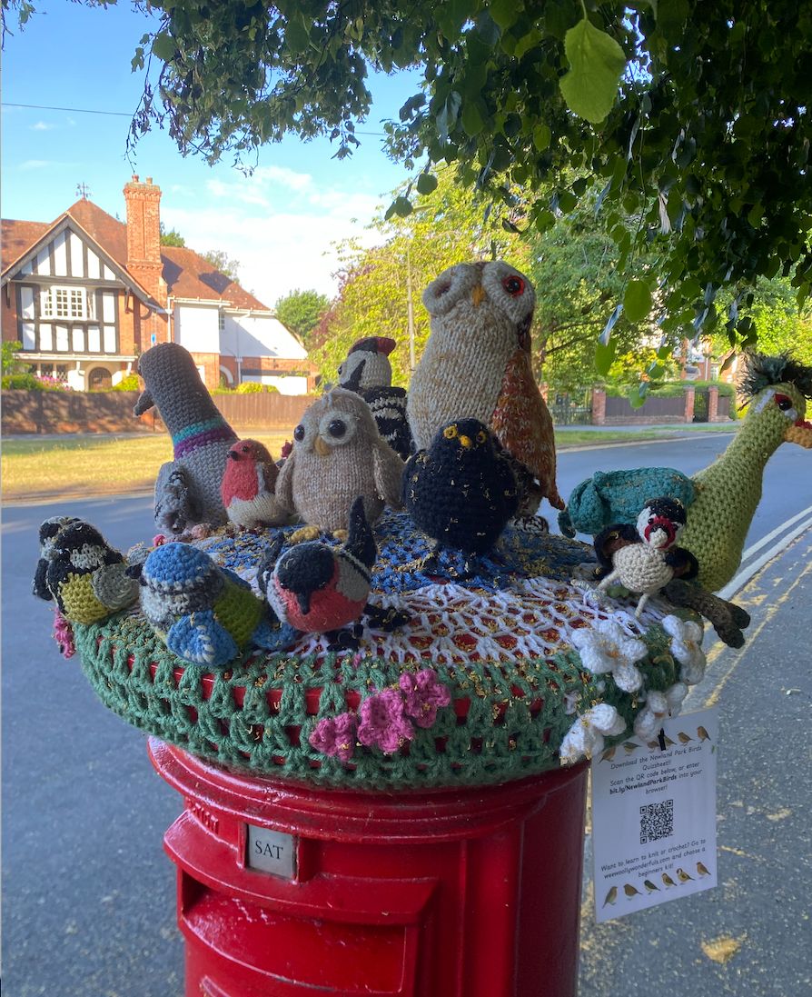 Knitted and crocheted group of birds topping a red postbox, with reference to NewlandParkBirds in a QR code