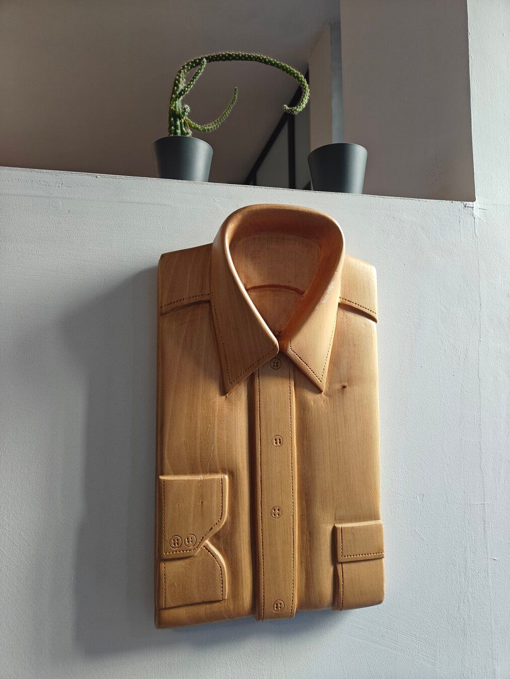A close-up photograph of a folded men's dress shirt skillfully carved from light-brown wood, complete with collar, button placket, pocket, and cuff details, mounted on a textured grey wall. Above the wooden carving, a small, curving cactus in a dark pot and another empty dark pot sit on a ledge. The wood grain is visible, and soft shadows are cast to the left.