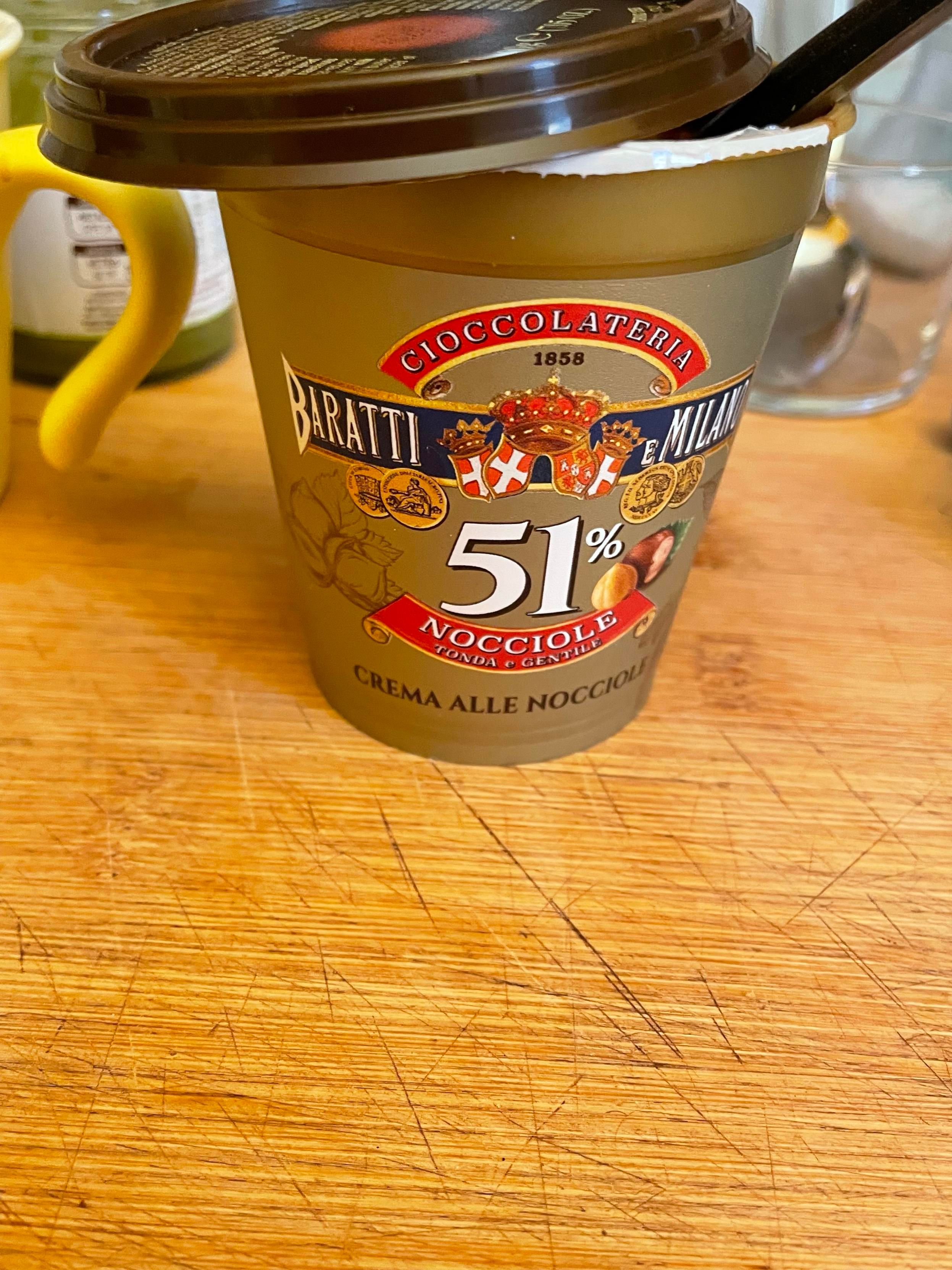L'immagine mostra una vaschetta di crema alla nocciola posizionata su un tagliere di legno. La vaschetta è contenuta in una scatola bianca e marrone con un coperchio trasparente. Sulla scatola si legge "CIOCCOLATERIA MILANO". Sotto questa scritta, si legge "51%". Sotto, si legge "NOCCIOLE". Sotto queste parole, c'è un logo con il disegno stilizzato di una testa. Sotto a questo, si legge "1858". L'intero design è in tonalità di marrone, bianco e oro. Il tagliere di legno ha una grana visibile ed è leggermente sgualcito.