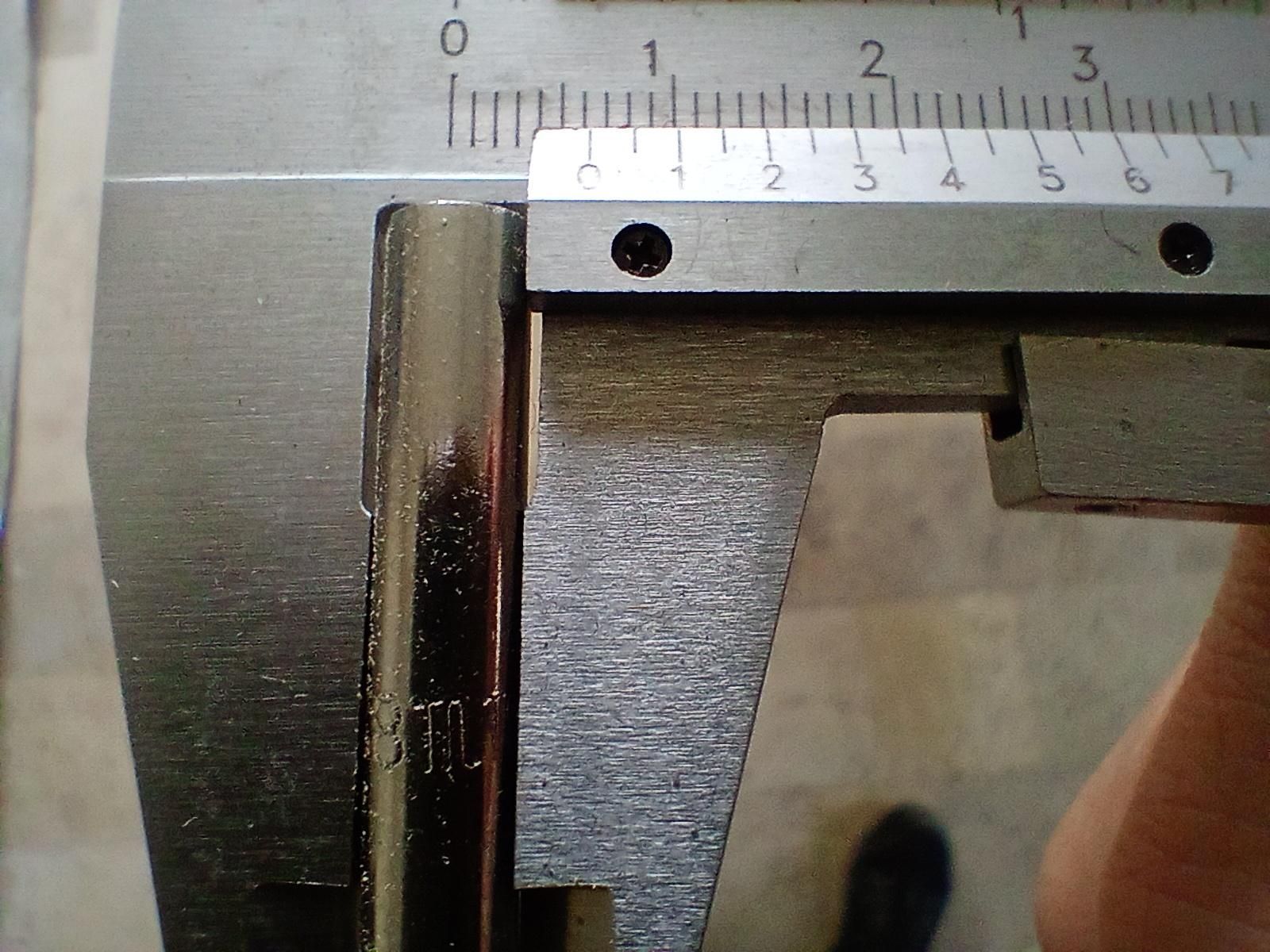 Un'inquadratura ravvicinata di un calibro metallico che misura un oggetto cilindrico. Il calibro presenta un corpo principale metallico con due fori circolari vicino al bordo superiore e una mandratura mobile. L'oggetto cilindrico a sinistra ha “311L” inciso sulla sua superficie. Sotto, una livella mostra marcature numeriche da 0 a 7 con piccole traccia di divisione. Una mano parzialmente visibile sul lato destro dell'immagine.