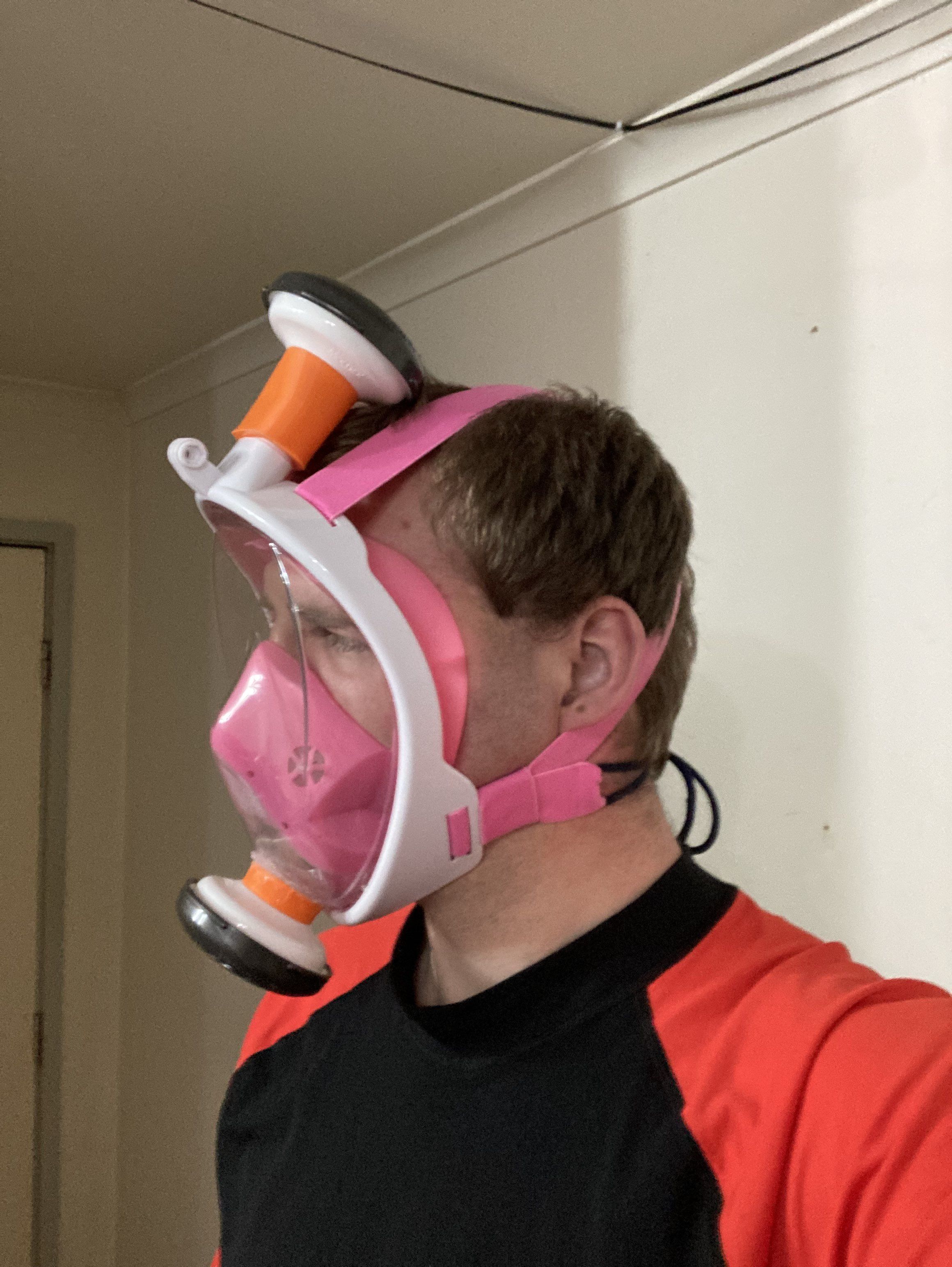 Upper body and head of a person (me) in side profile wearing a white and pink snorkeling mask. Orange 3d printed spacers are attached to the breathing ports of the mask at the top and bottom and have respirator particle filters attached to them. The ensemble forms a DIY respirator.