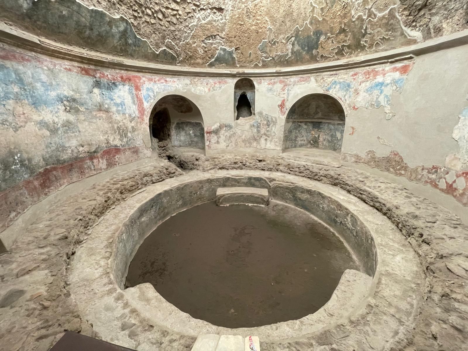 Piscina delle terme. Una vasca circolare centrale, con sopra due nicchie speculari e una nicchia più piccola in mezzo, leggermente rialzata. Le pareti, circolari, hanno resti di intonaco colorato