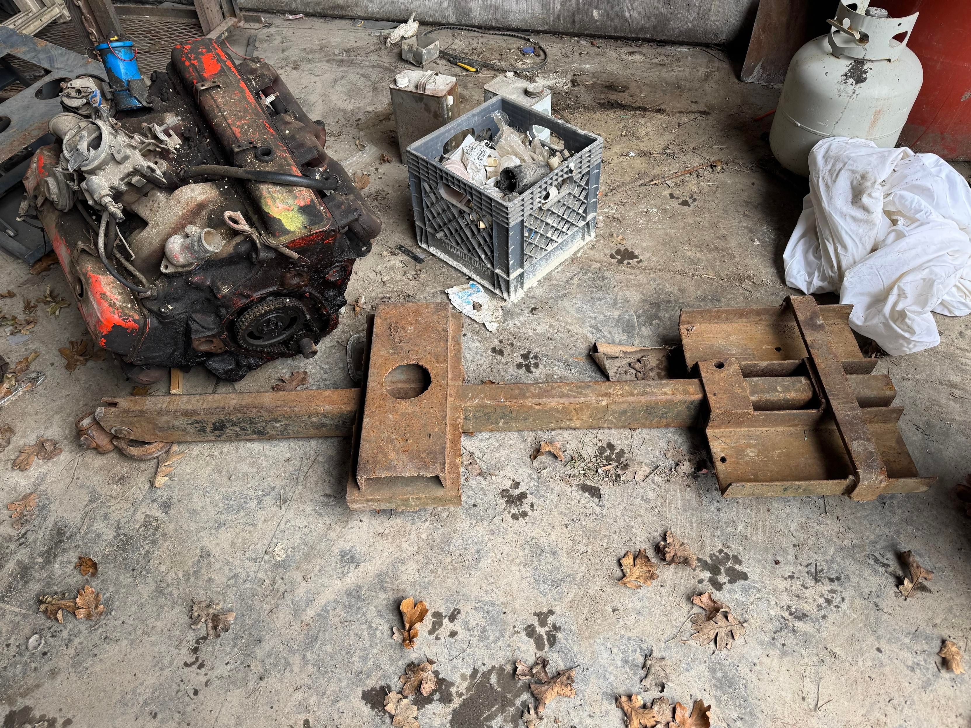 An industrial setting with a disassembled engine in the foreground and a large, heavy metal tool or fixture lying nearby. A plastic crate filled with miscellaneous items is also visible, along with a propane tank and a pile of white fabric. The floor is scattered with junk 