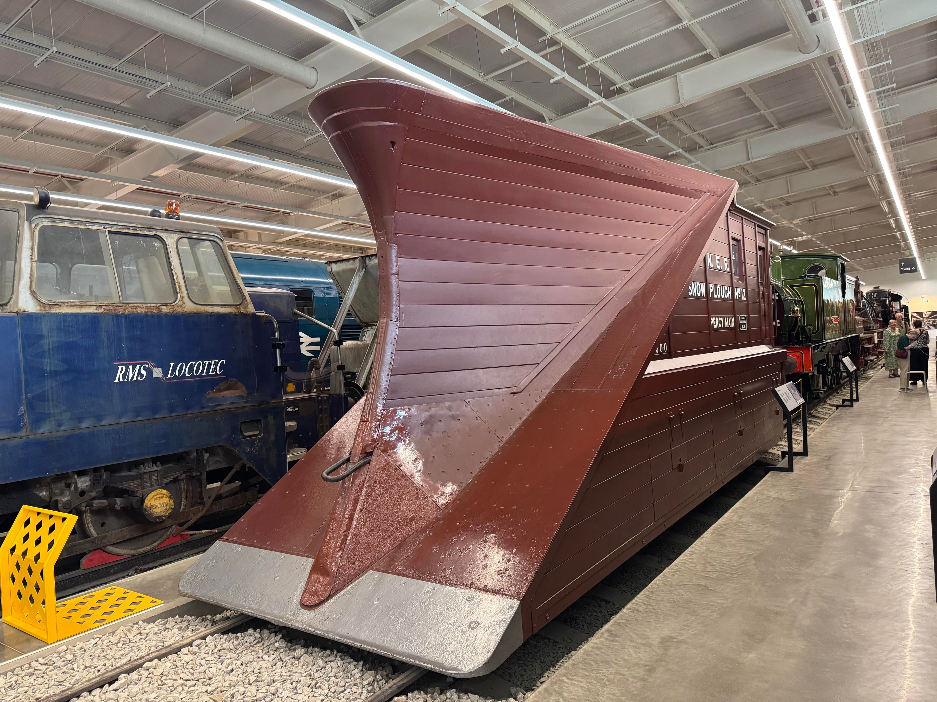 A 3/4 side view of a big reddy-brown painted wooden snowplough seen from the front.