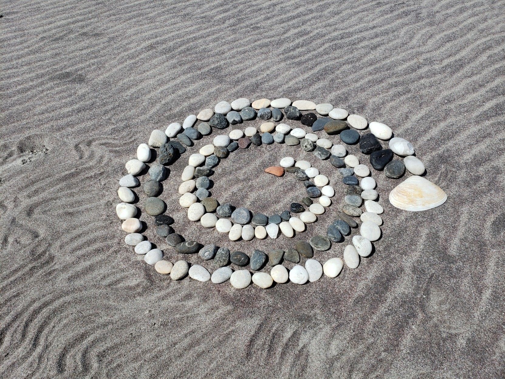 A spiral made of graduated size light and dark stones, starting at a orange-red stone and spiralling to a clamshell. It's on top of dark red sand (garnet) with wind and finger-raked ripples.