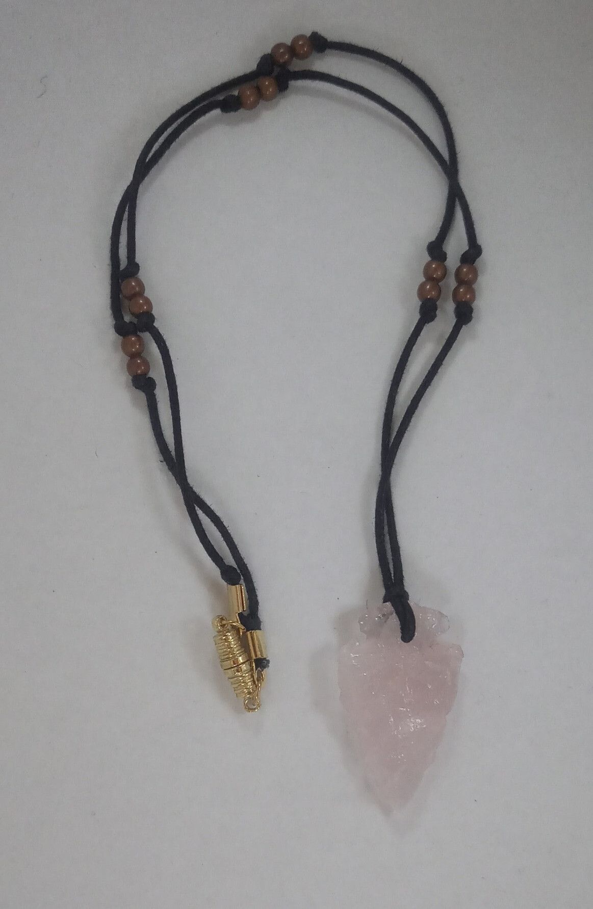 A clear rose (pink) quartz arrowhead pendant, on a white background; the arrowhead has been knapped, so it has lots of rough, chipped-looking edges. It's strung on a black cord, with pairs of small copper-finished sphere beads at short intervals, and a gold-finished oblong magnetic clasp. The cord has been looped through a hole in the top of the arrowhead to hold it. 
