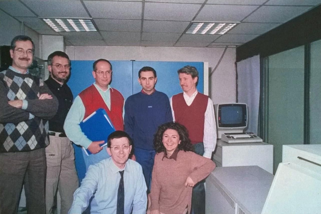 Un gruppo di sette persone posa sorridendo in un ufficio tecnico. Sei sono in piedi e una è inginocchiata davanti. Sono presenti uomini e una donna, vestiti in modo informale da lavoro. Sullo sfondo si vedono armadi tecnici blu e, a destra, un vecchio computer con monitor CRT e tastiera. L’ambiente ha luci al neon e un aspetto da sala macchine informatica degli anni ’80-’90.