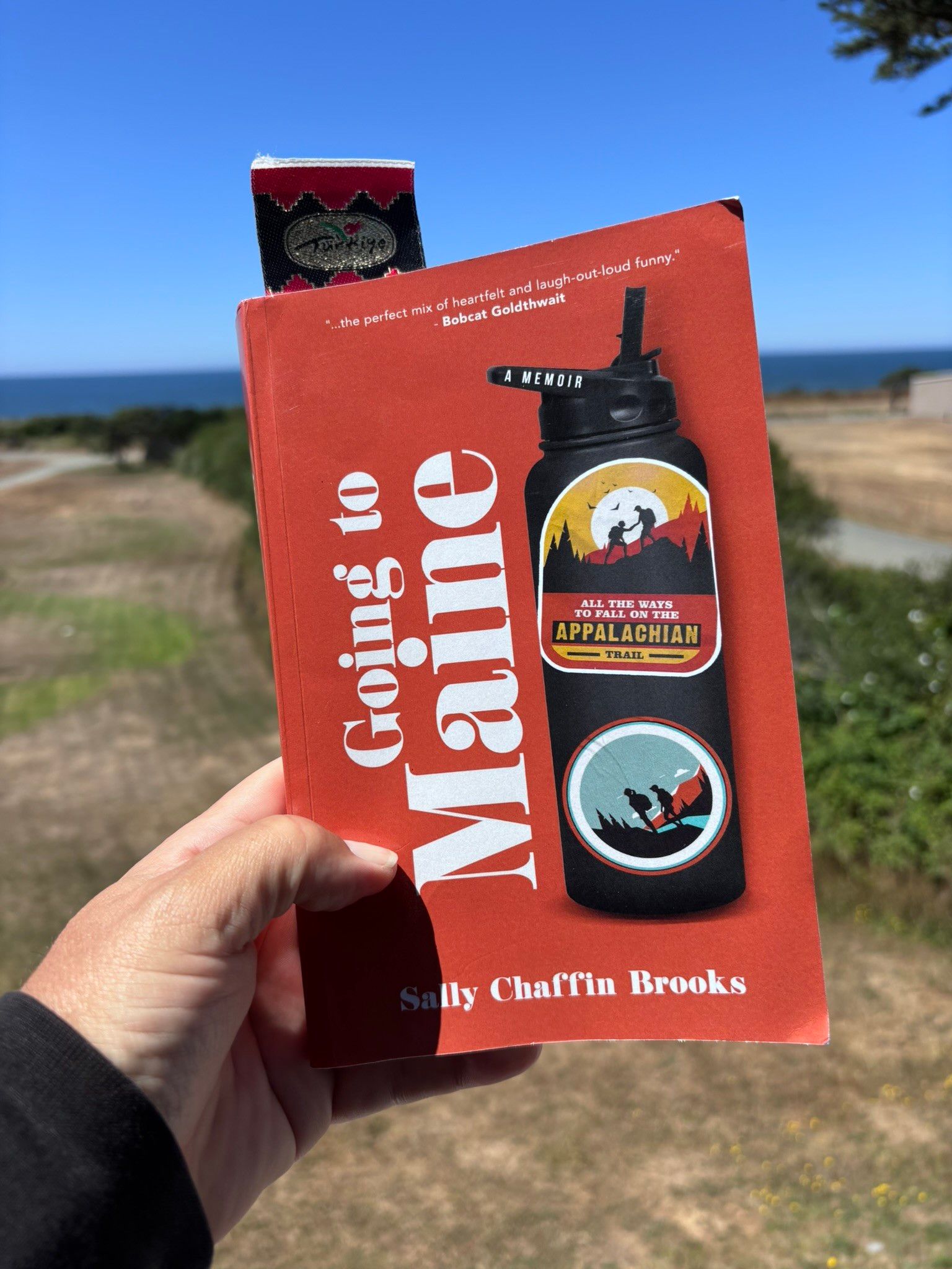 Hand holding the book "Going to Maine" by Sally Chaffin Brooks outdoors with a scenic background of fields, trees, and a body of water under a clear blue sky.