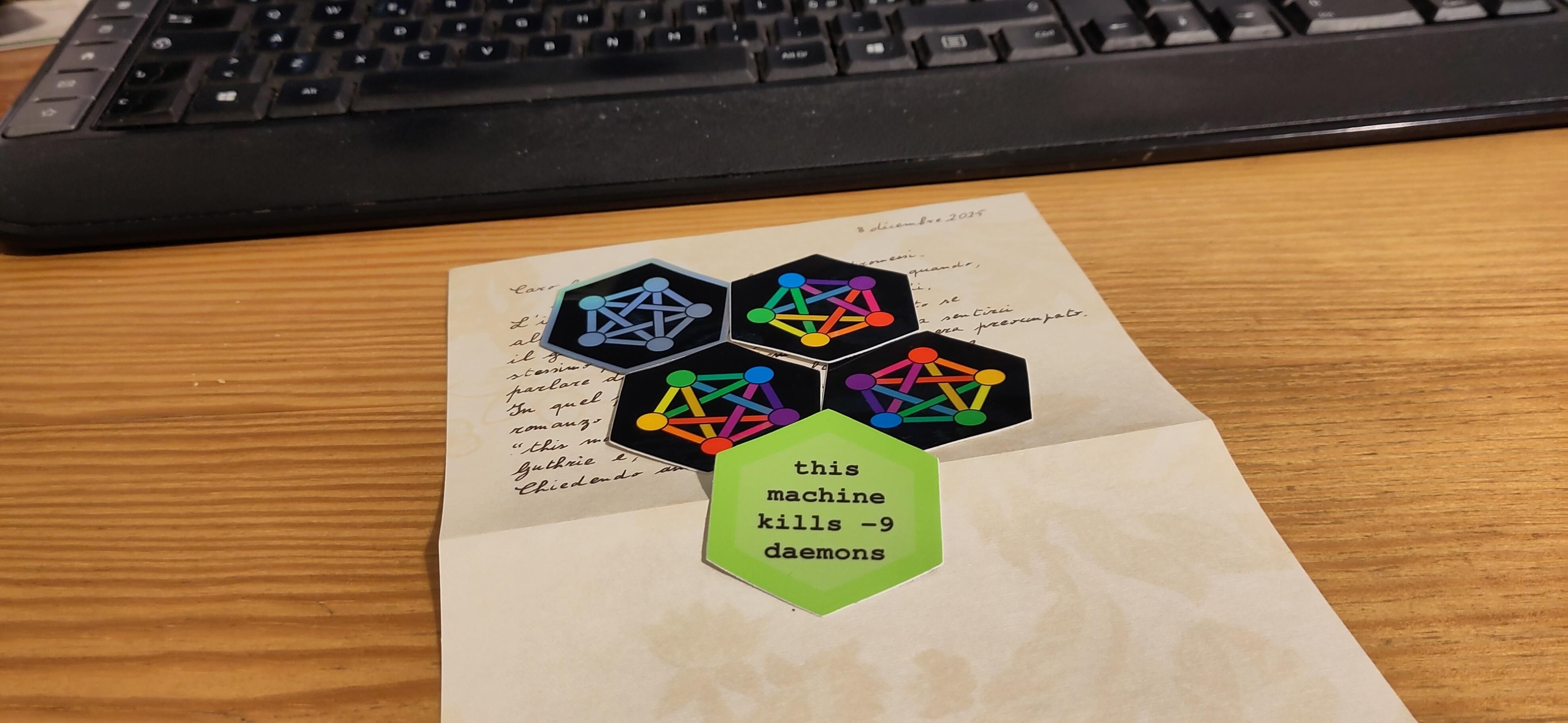 una lettera scritta a mano poggiata su una scrivania di legno davanti una tastiera, con degli adesivi esagonali con il logo del fediverso sopra, ed uno uno che recita "this machine kills -9 daemons"