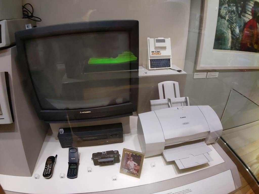 Eine Vitrine mit einem Fernseher, einem Tintenstrahldrucker, einer kompaktkamera und zwei Handys.