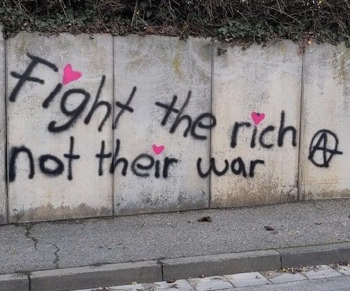 Scritta su un muro: "Fight the rich non their war" con una A cerchiata.